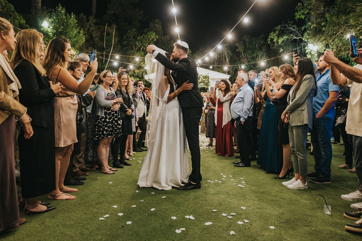 Couple sharing their first dance outdoors under string lights as wedding guests watch from both sides.
