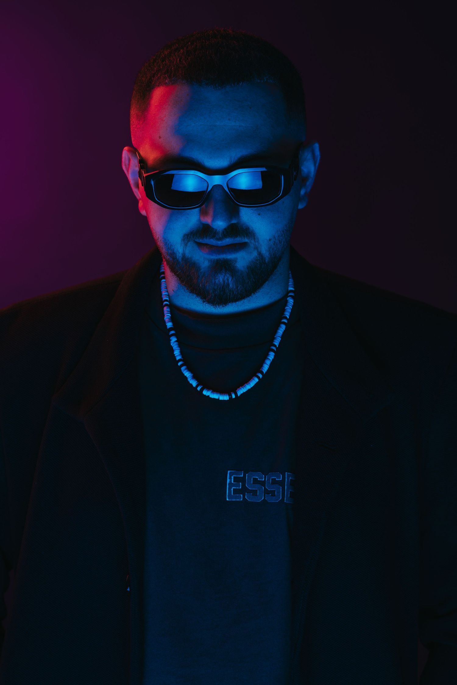 Neon-lit portrait with blue and purple lighting of person wearing sunglasses and chain necklace.