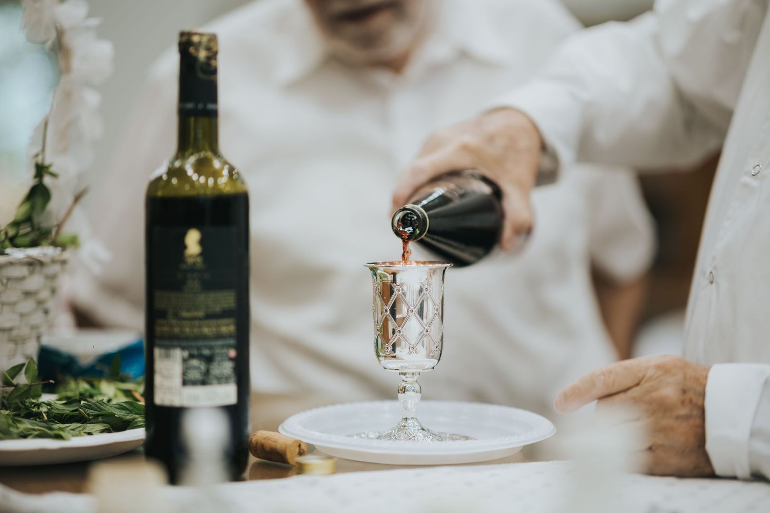 Hands pouring red wine into a crystal glass against a white tablecloth background.