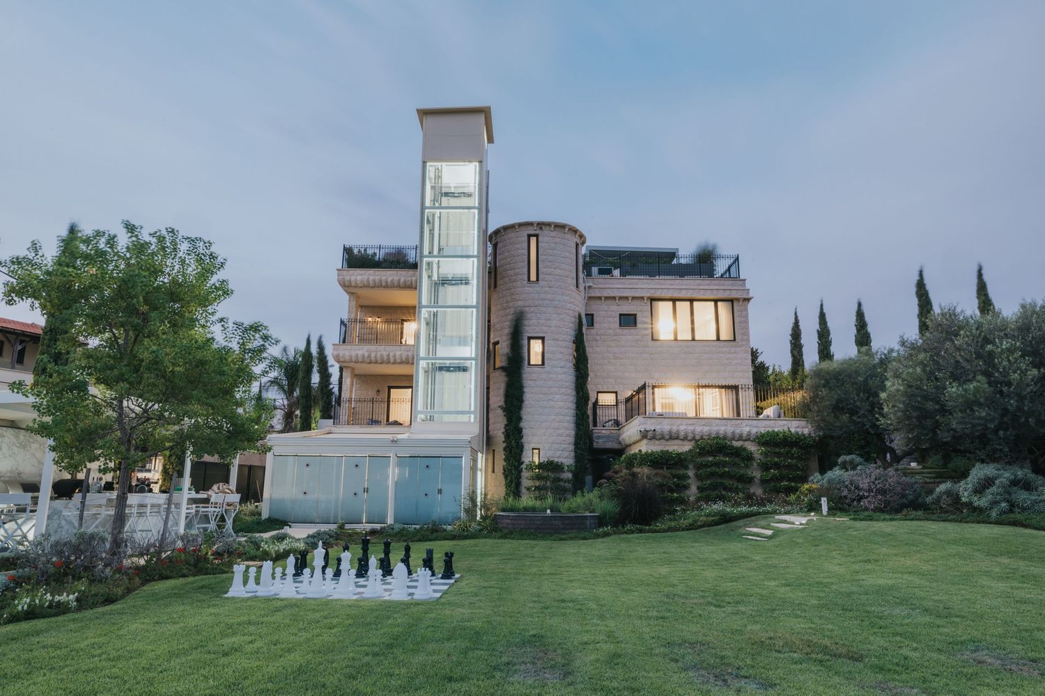 Contemporary multi-level villa with glass elevator tower and landscaped gardens at dusk.