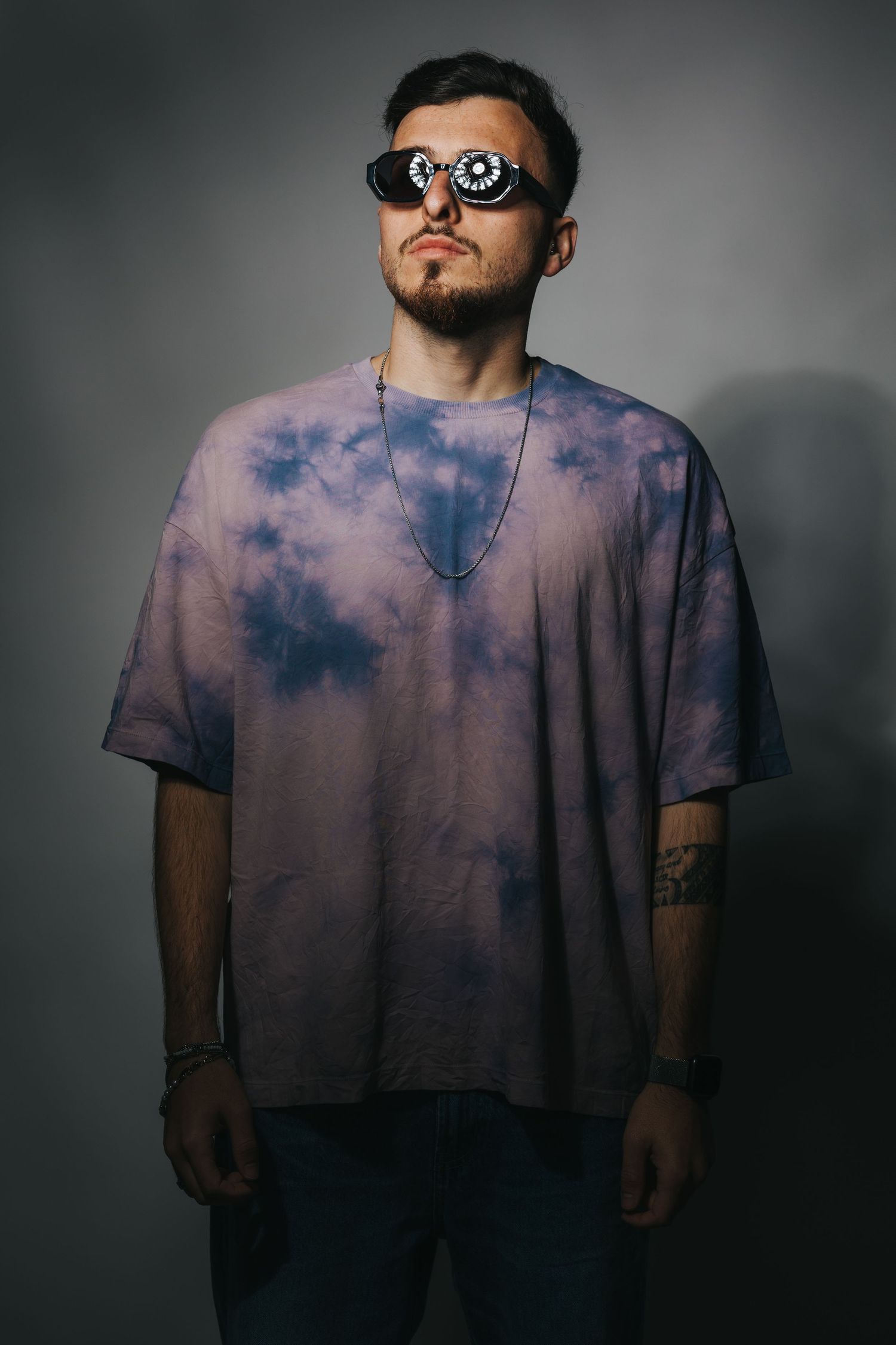 Person wearing tie-dye shirt and sunglasses poses against grey backdrop.