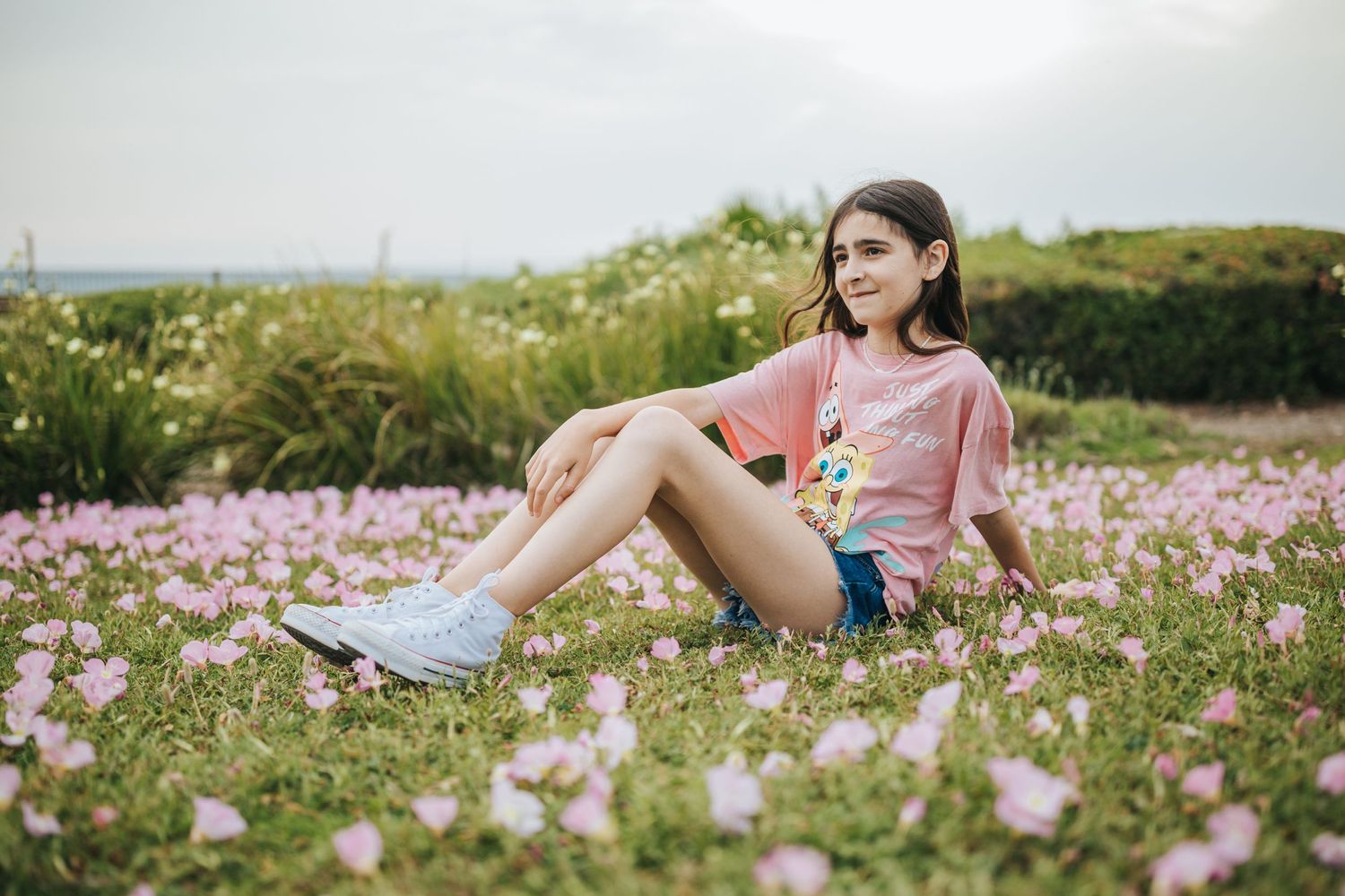 Person sitting in a field of pink cosmos flowers wearing pink top and denim shorts on a sunny day.