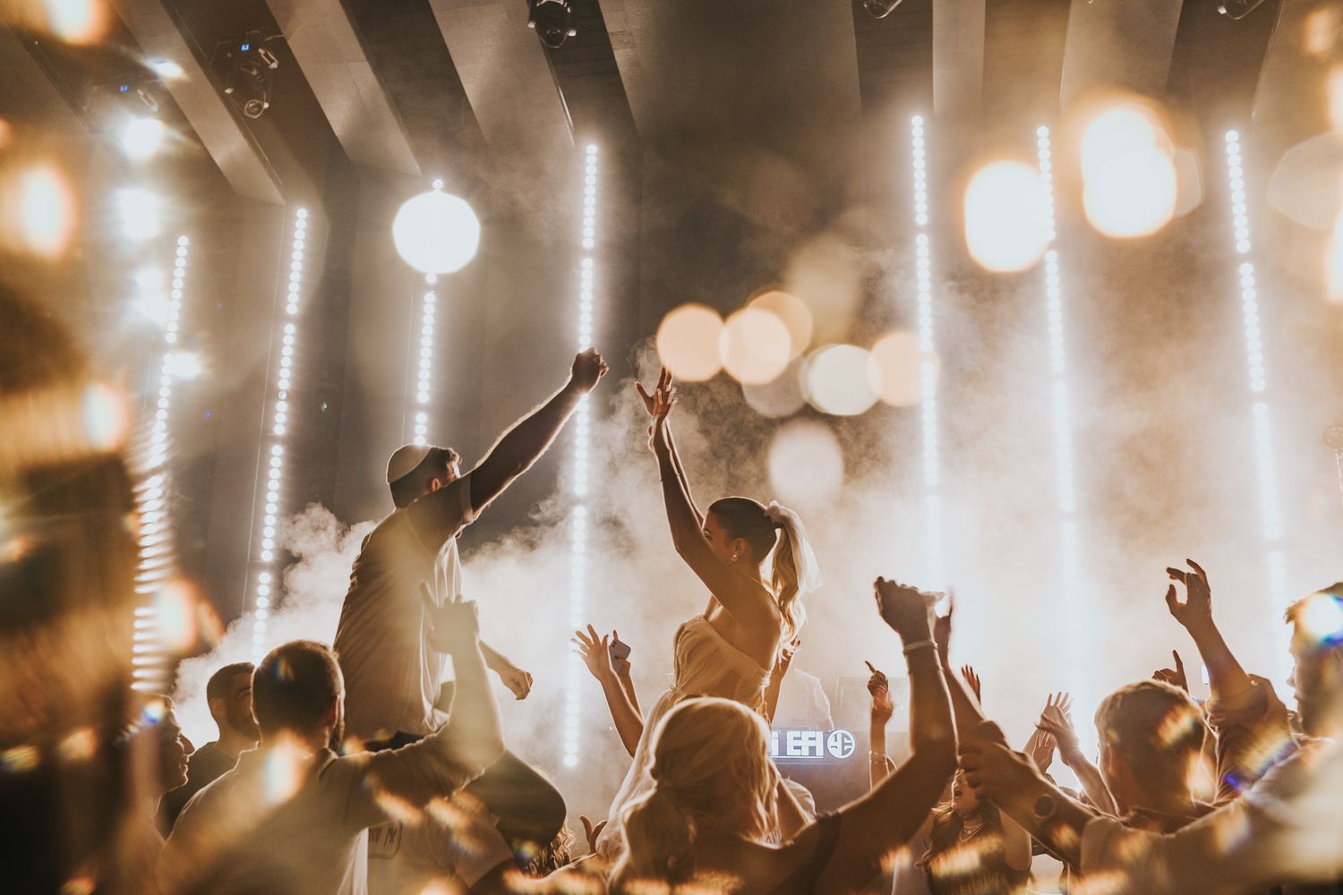 Concert crowd silhouettes with raised arms against bright stage lights and bokeh effects.
