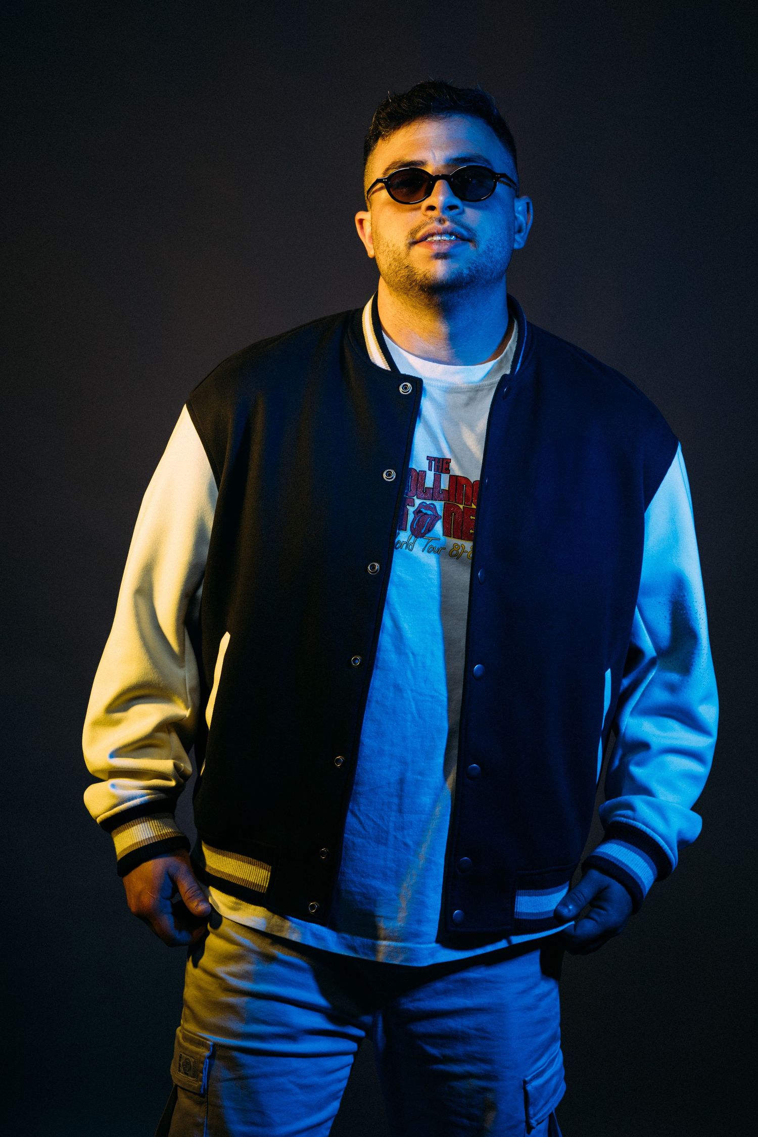 Modern artist poses in a varsity jacket and sunglasses against dark background with dramatic lighting.