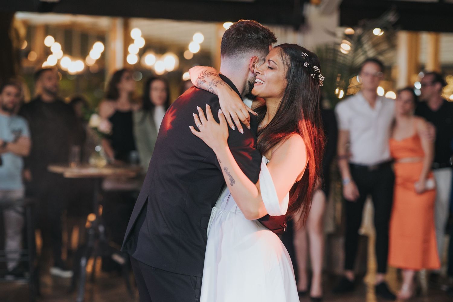 Romantic couple dancing intimately at evening reception with string lights and guests in background.