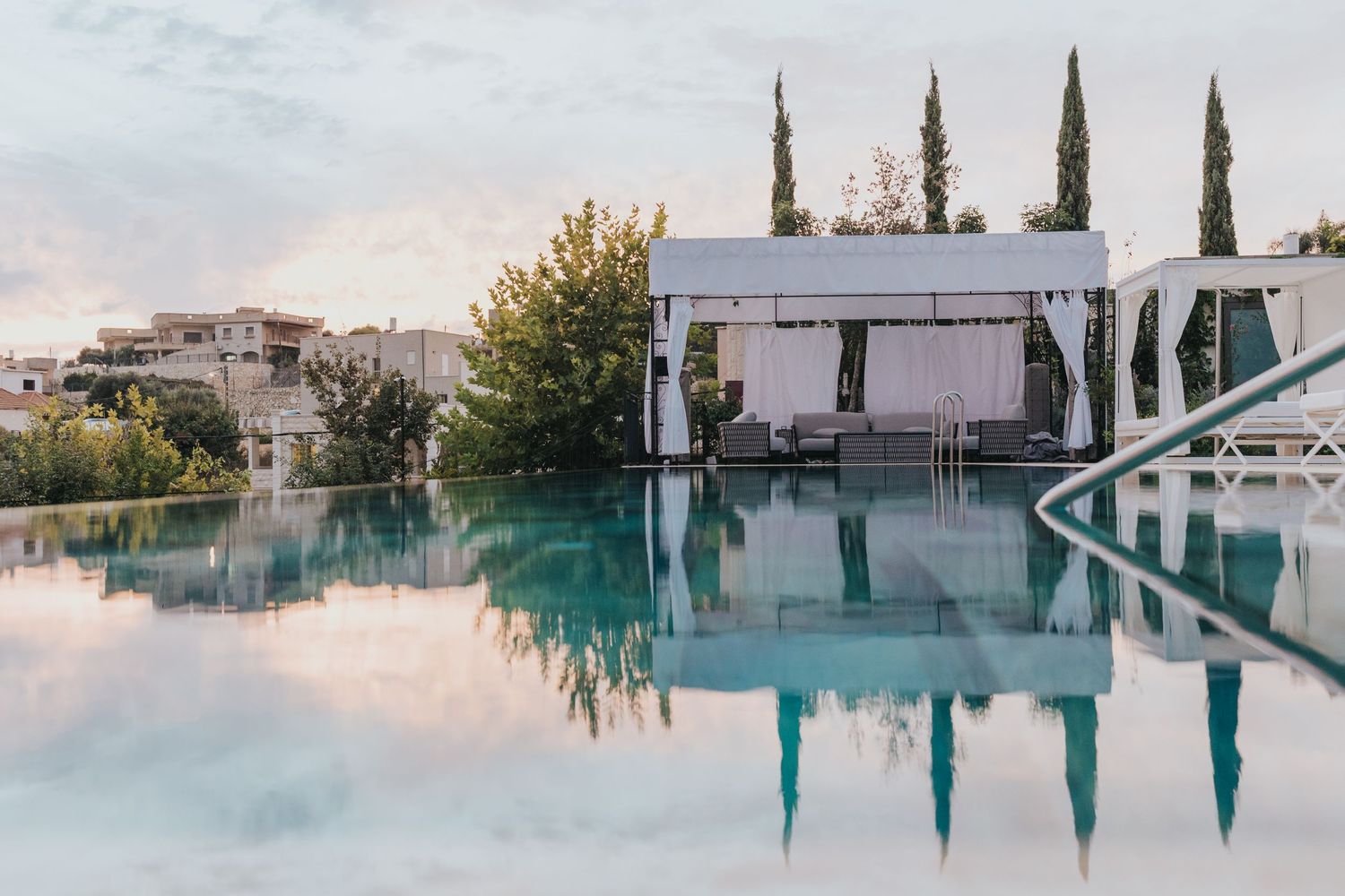 Luxurious infinity pool with white cabana and cypress trees at sunset.