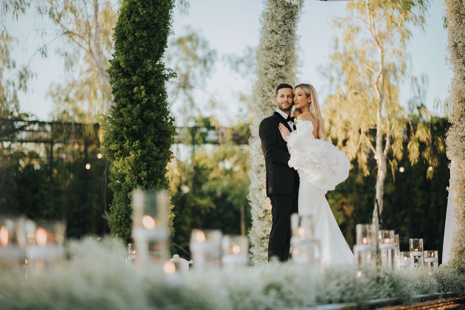 Couple in formal wedding attire standing together under greenery at sunset.