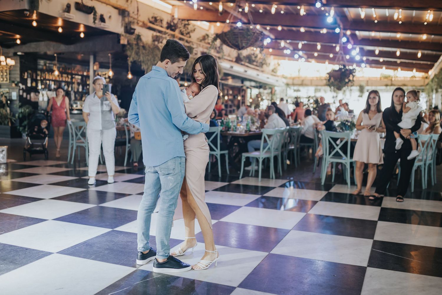 Couple dancing on checkered floor with string lights and guests in background.