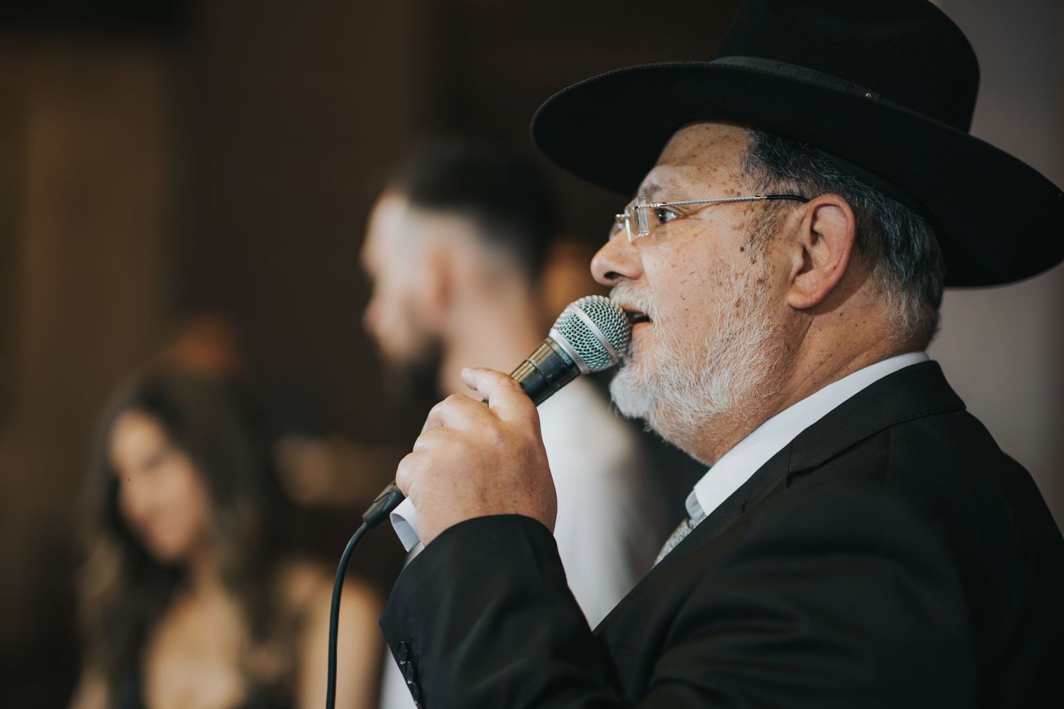 Speaker in traditional black attire and hat using microphone during ceremonial event.