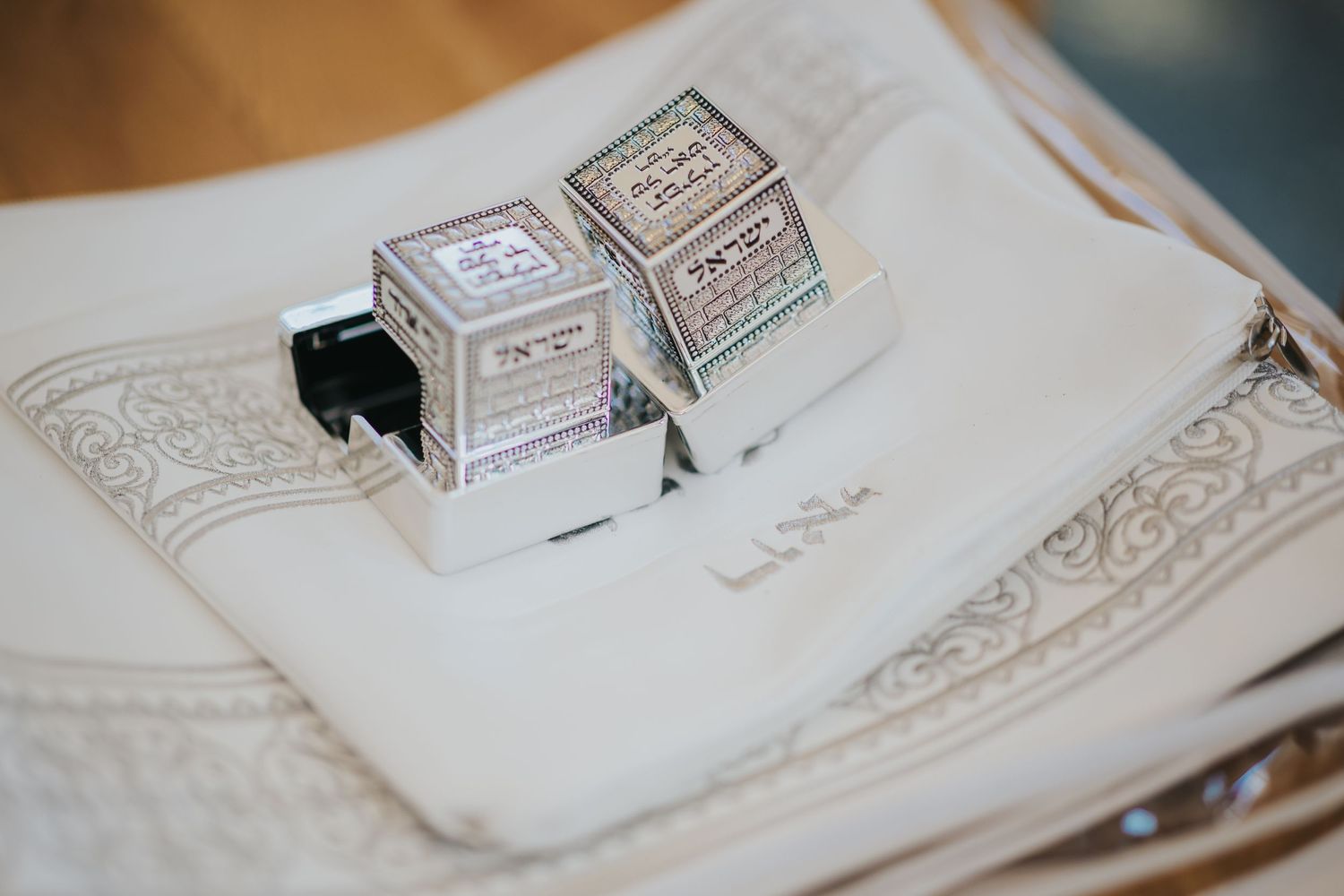 Silver ritual tefillin boxes resting on an ornately decorated white fabric.