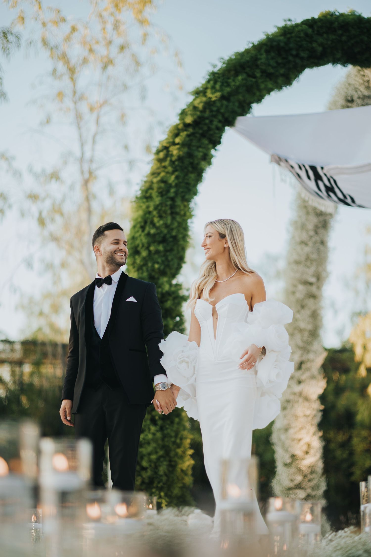 Elegant wedding couple poses under a lush green archway at an outdoor ceremony.