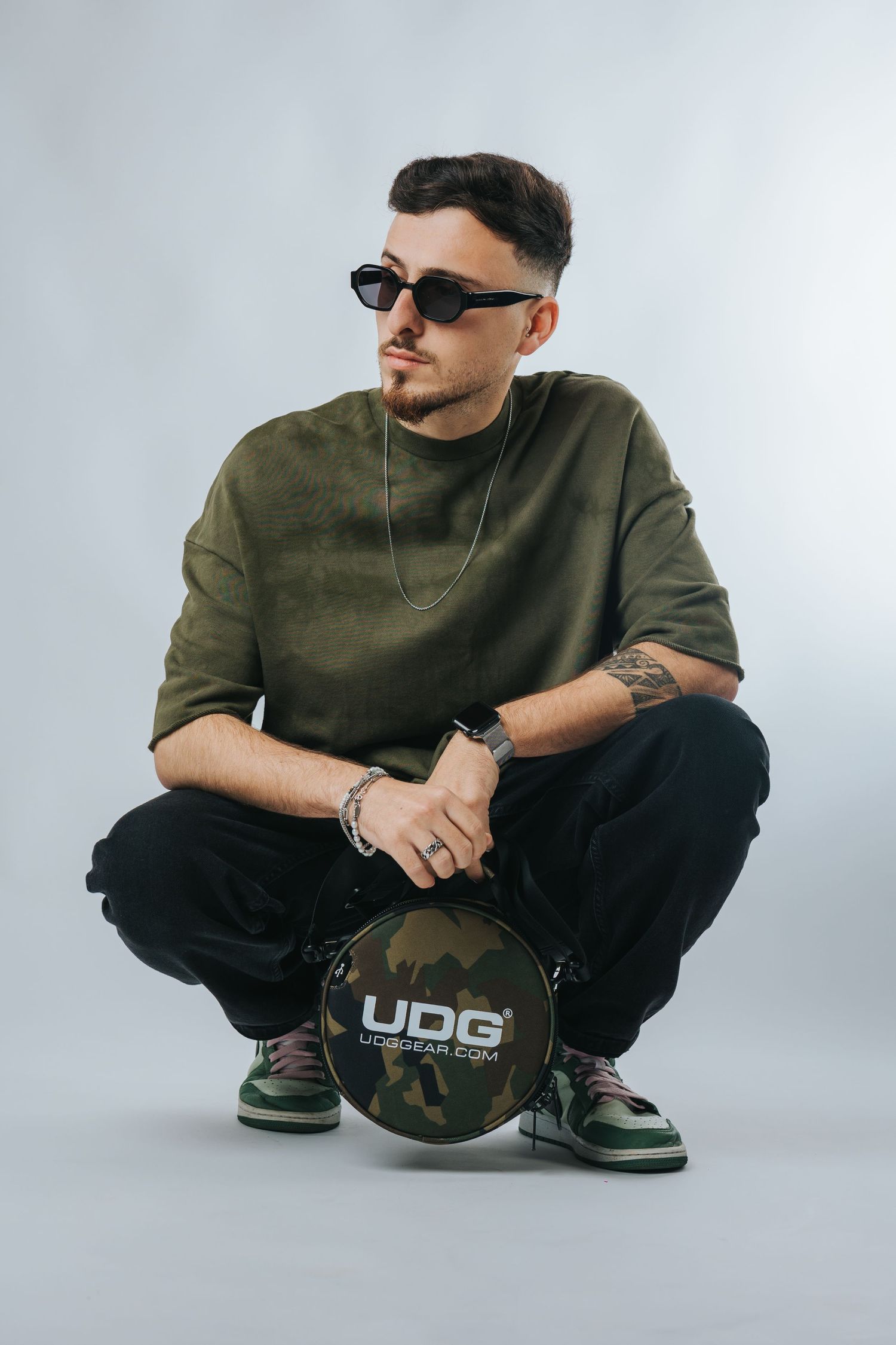 A DJ crouches down with sunglasses while holding a UDG branded disc against a white background.