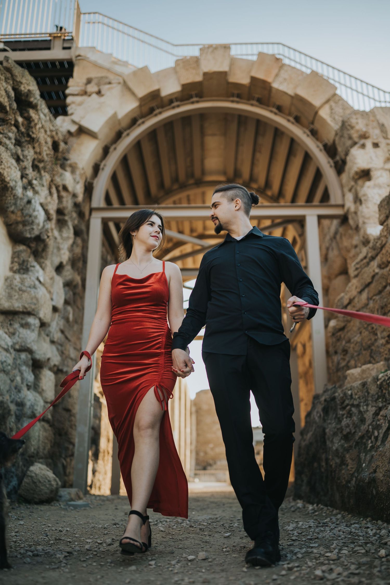 Couple walking through stone archway, woman in red dress and man in black attire.