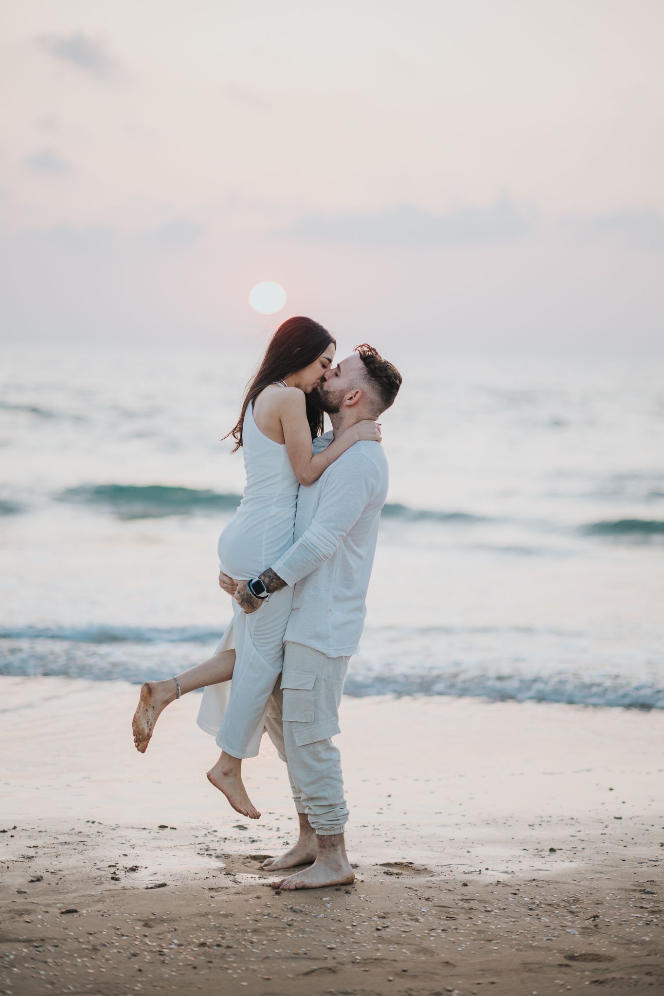 Couple embracing on beach at sunset with one person lifted off the ground in a romantic pose.