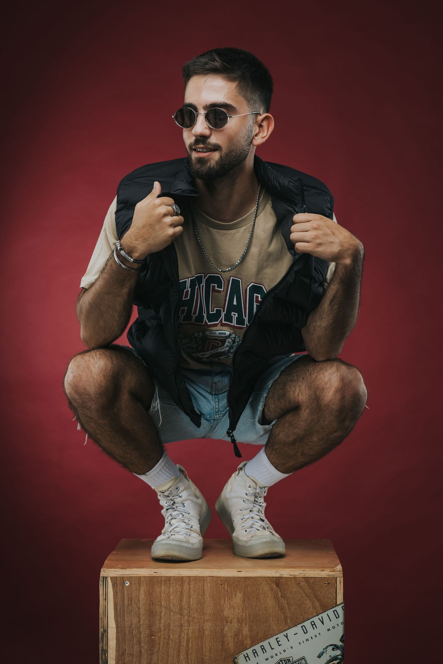 Urban fashion portrait of a person in sunglasses and black vest crouching on a wooden platform against a red background.