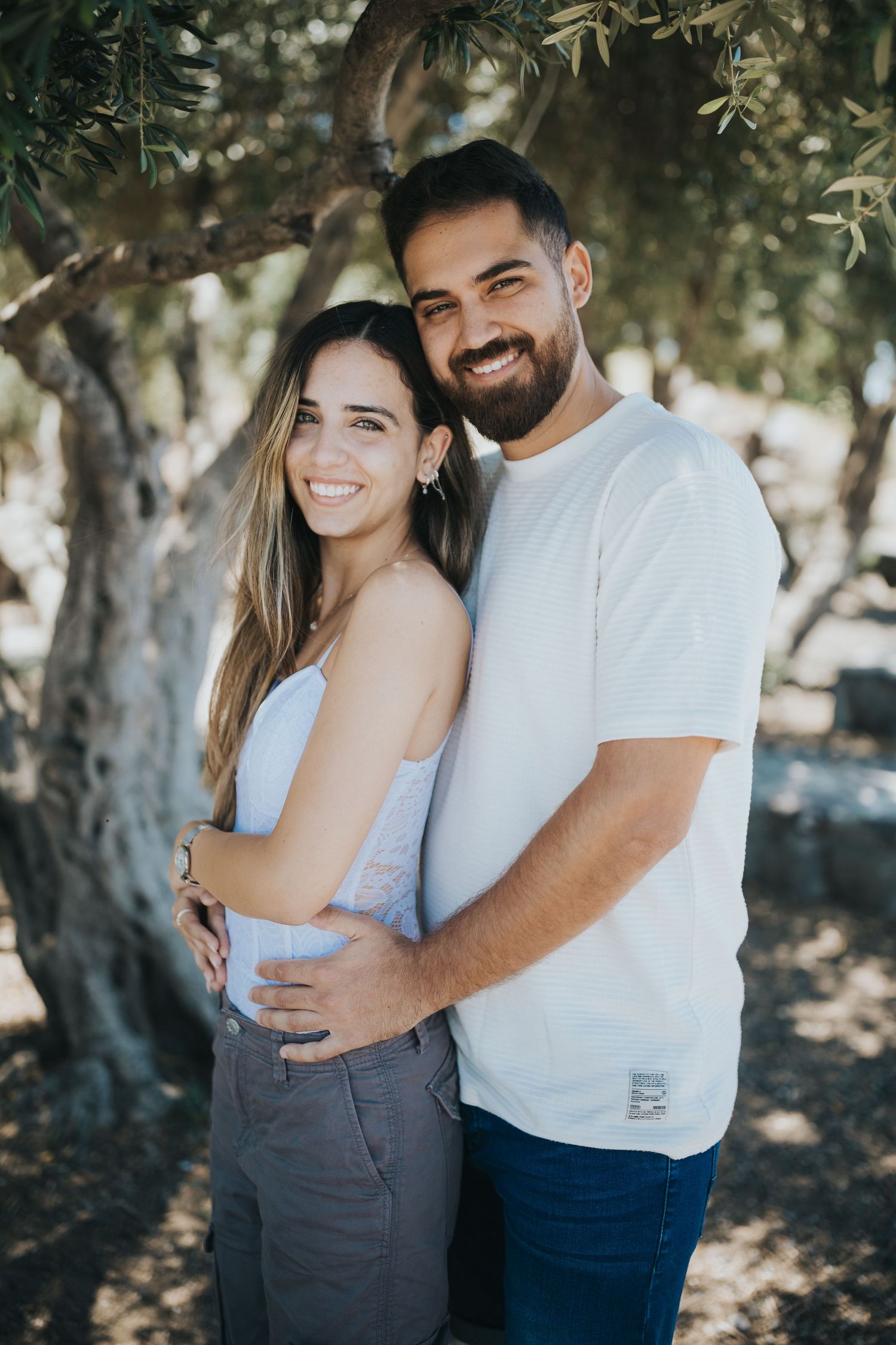 Couple in casual white outfits embracing under olive trees in natural outdoor setting.