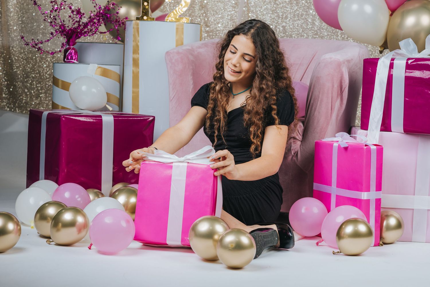 Festive scene with pink gift boxes, gold balloons and decorations around plush chair.