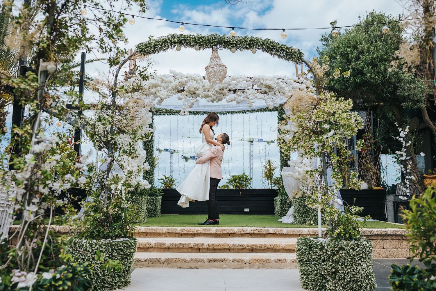 Newlyweds share a romantic moment under a floral arch in a garden setting.