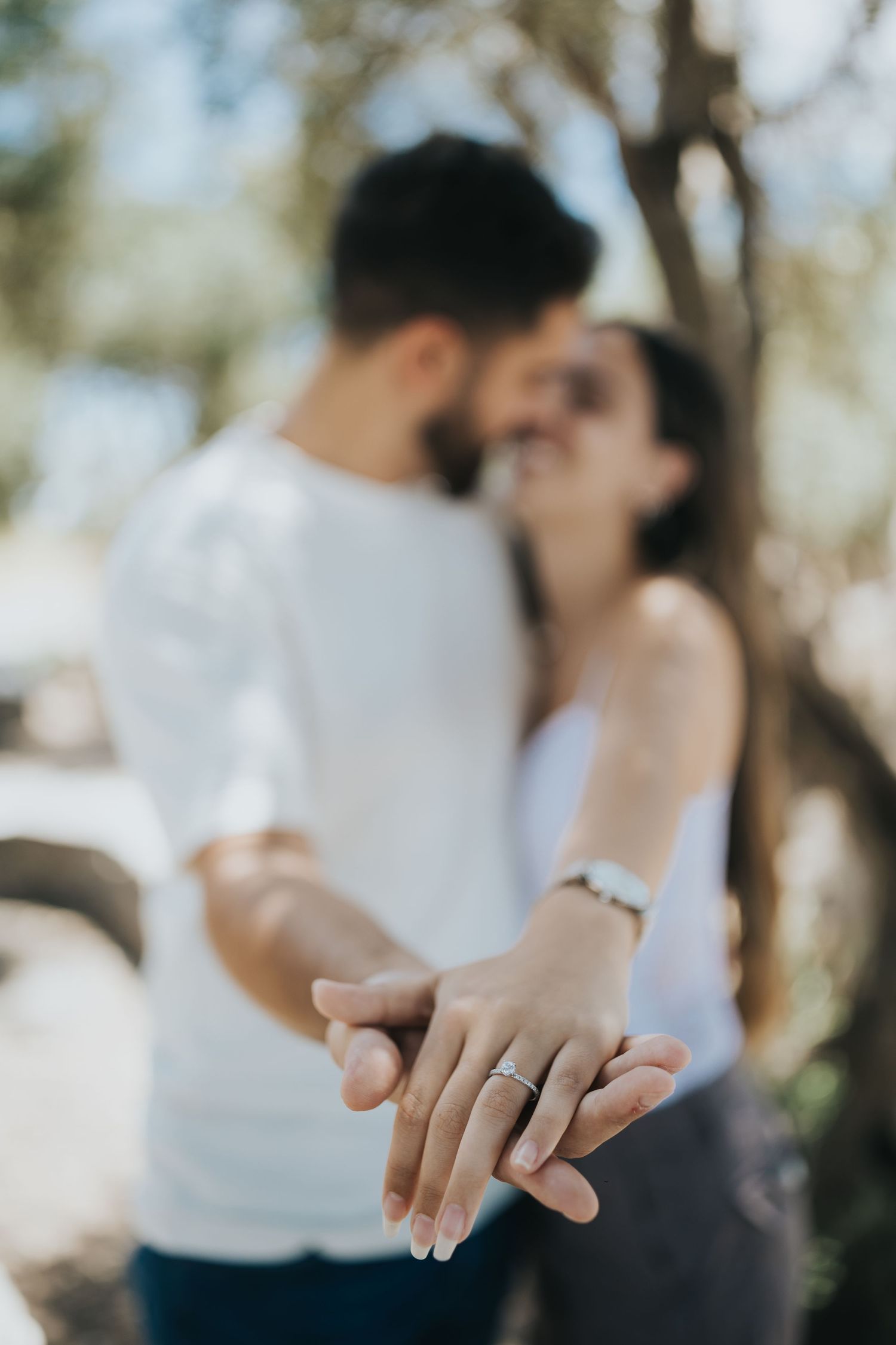 Close-up shot of hands holding with engagement ring in focus and blurred couple in background.