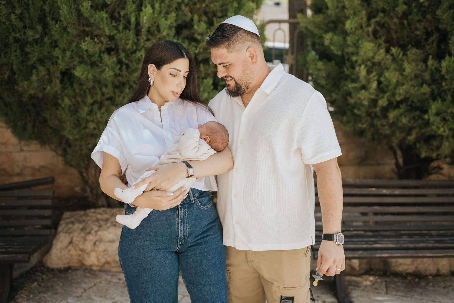 A couple stands outdoors holding their baby while enjoying a sunny day among green foliage.