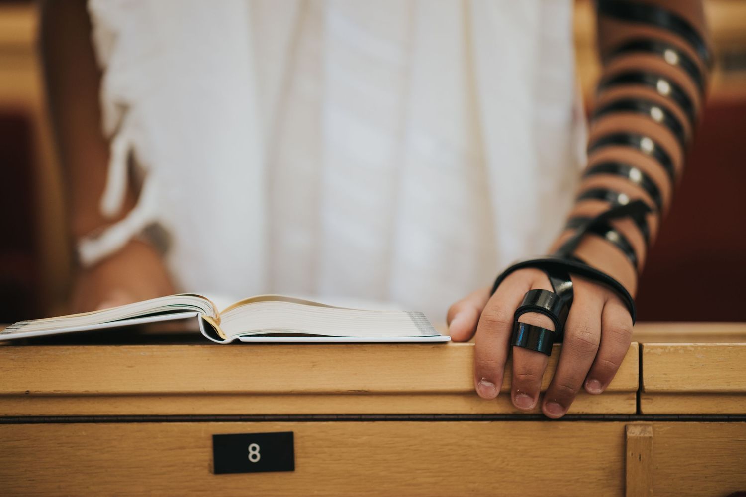 Hand with tefillin strap resting on an open book with a number 8 visible.