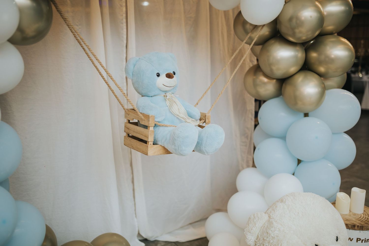 Blue teddy bear decoration on a swing with gold and white balloon arch display.