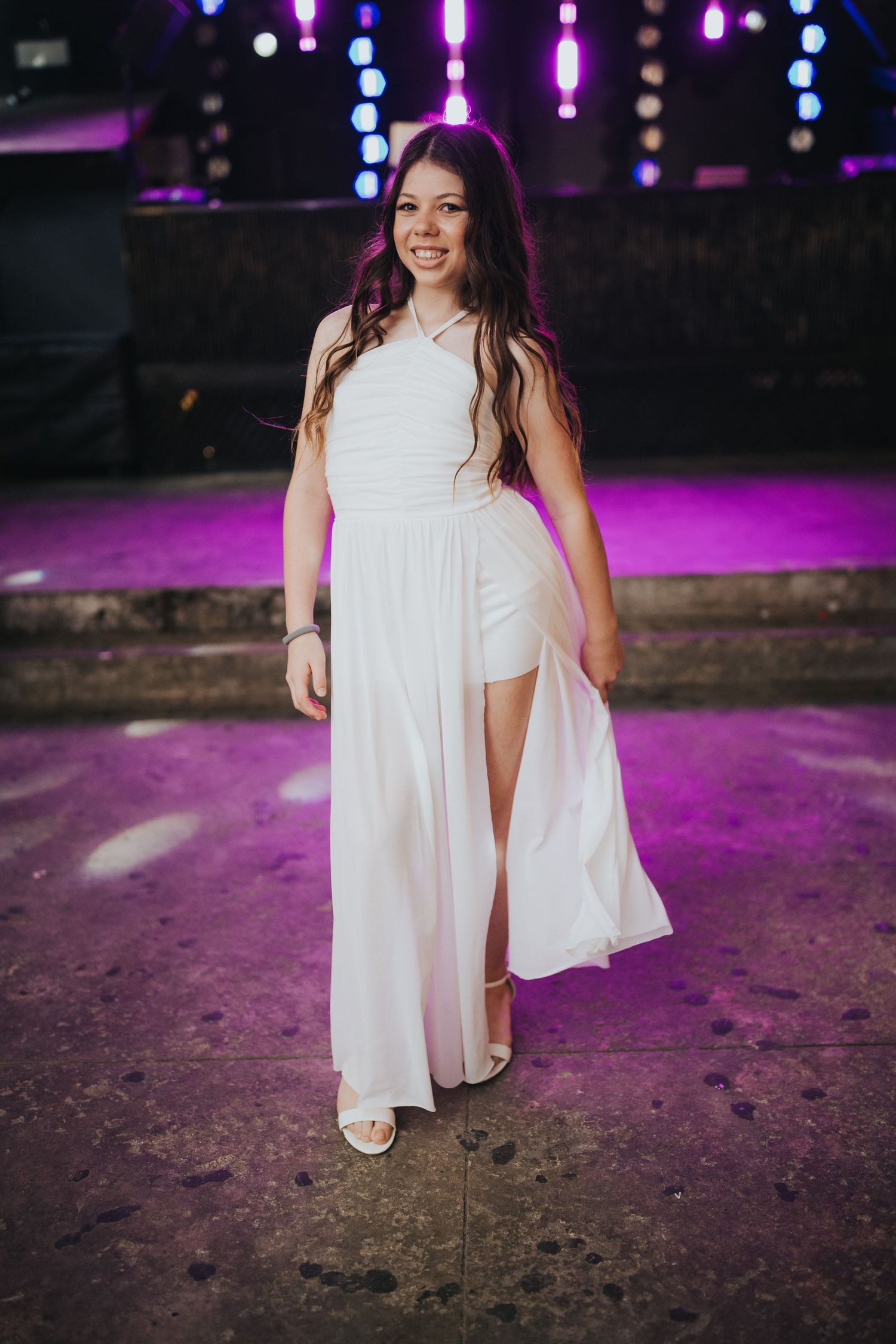 A figure in a flowing white dress poses against a purple-lit background with hanging lights.
