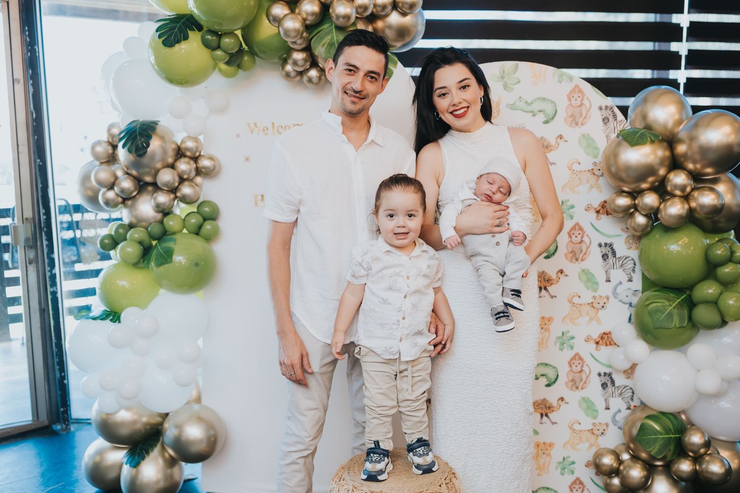 A family poses together against a backdrop decorated with green and gold balloon garlands for a special celebration.
