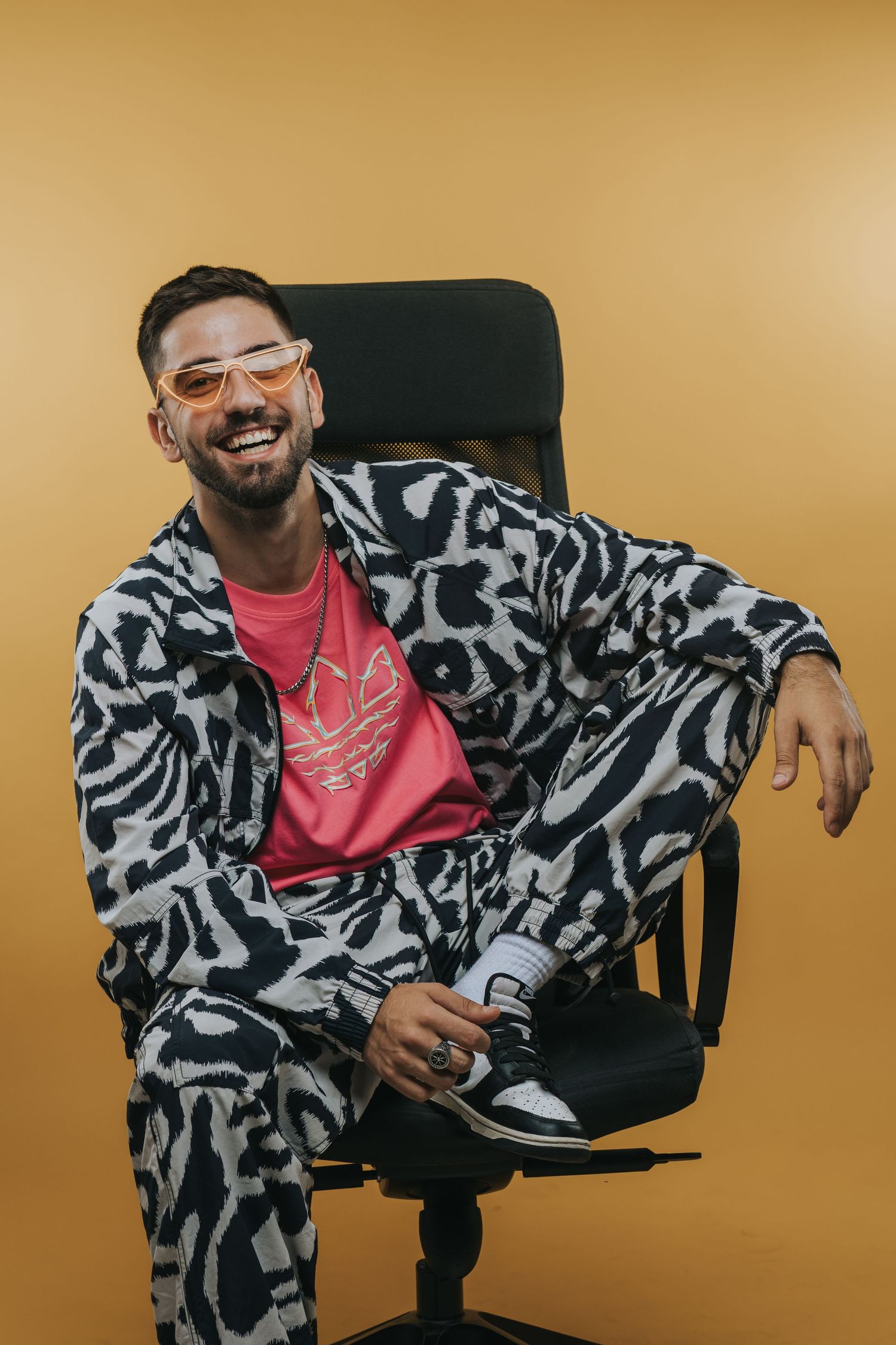 Person in zebra print outfit and pink shirt sits casually in office chair against golden background.