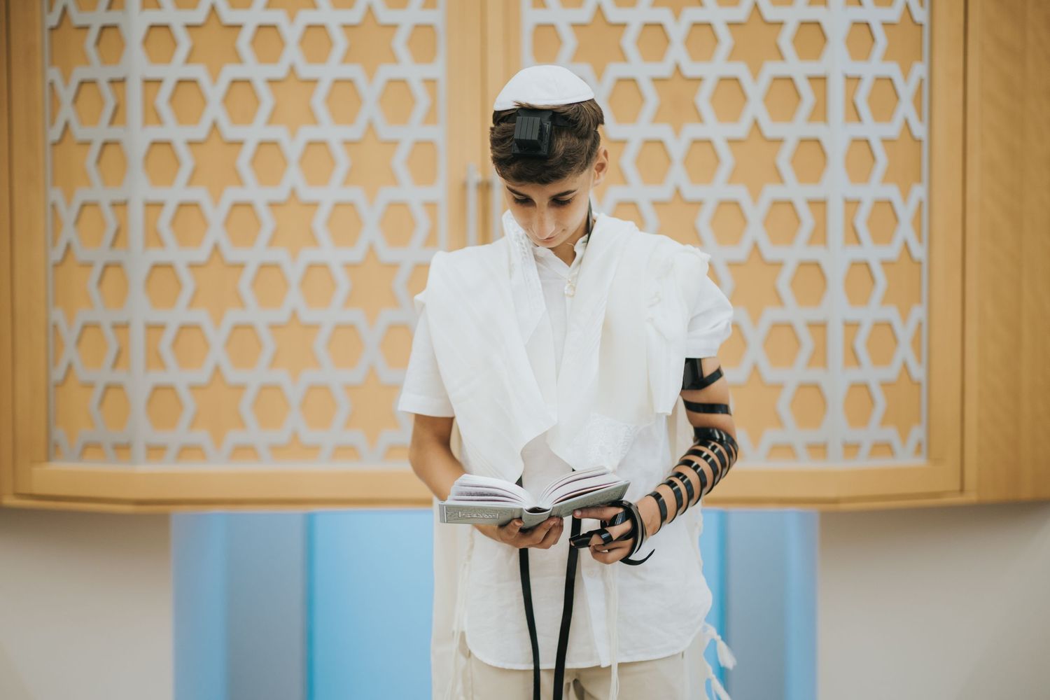 A person reads from religious texts while wearing prayer garments in front of a decorative wall.