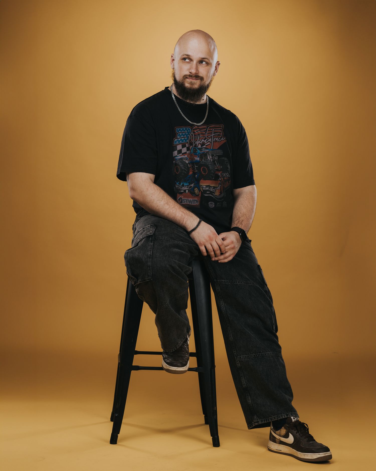 Studio portrait of someone in black t-shirt and cargo pants sitting on a stool against orange background.