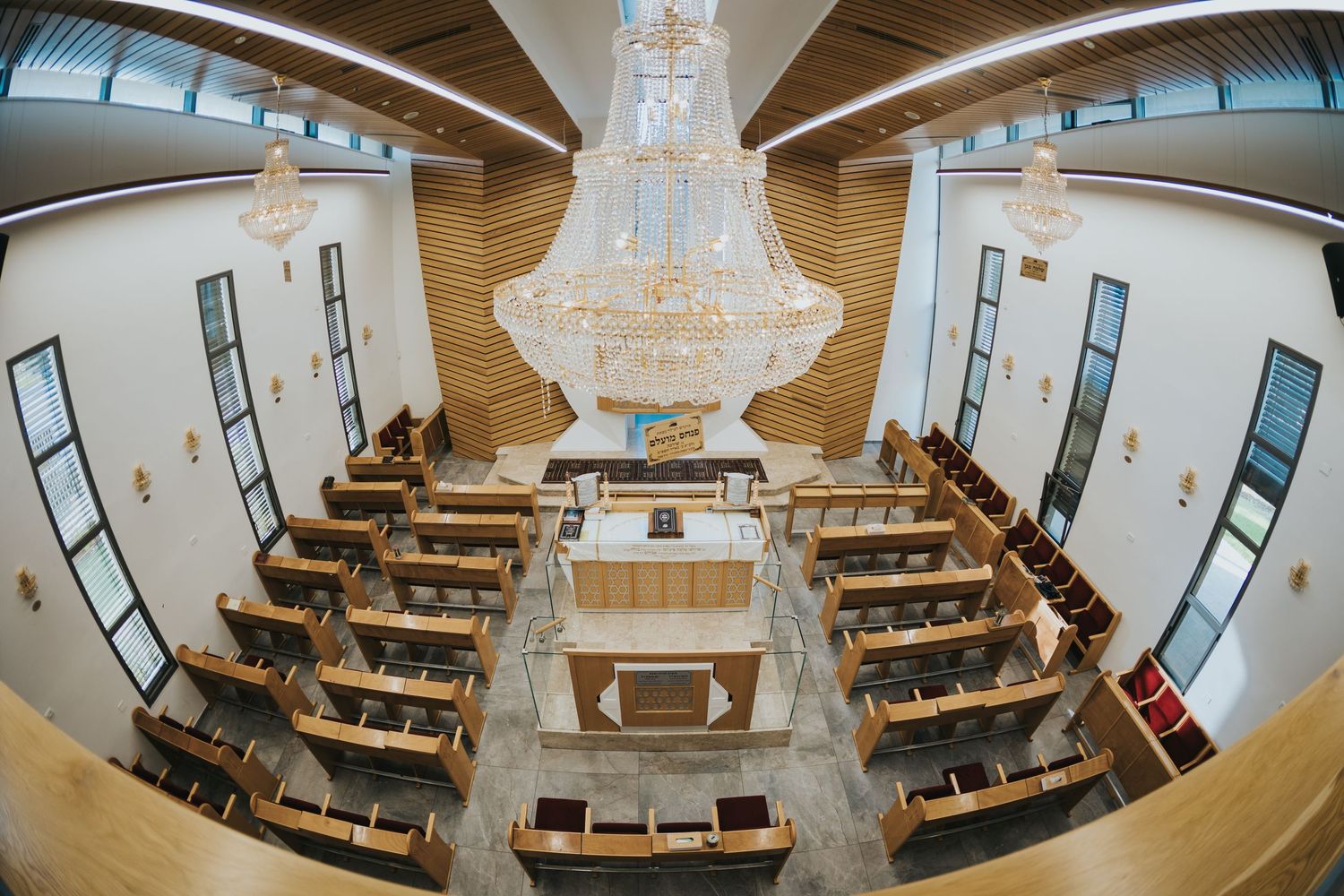 Modern synagogue interior with wooden pews and a grand chandelier viewed through a fish-eye lens.