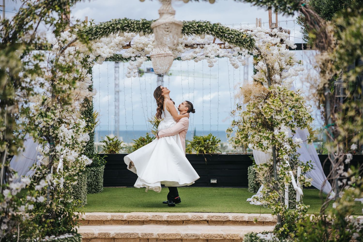 Bride in white dress dancing under a floral arch with ocean view backdrop.