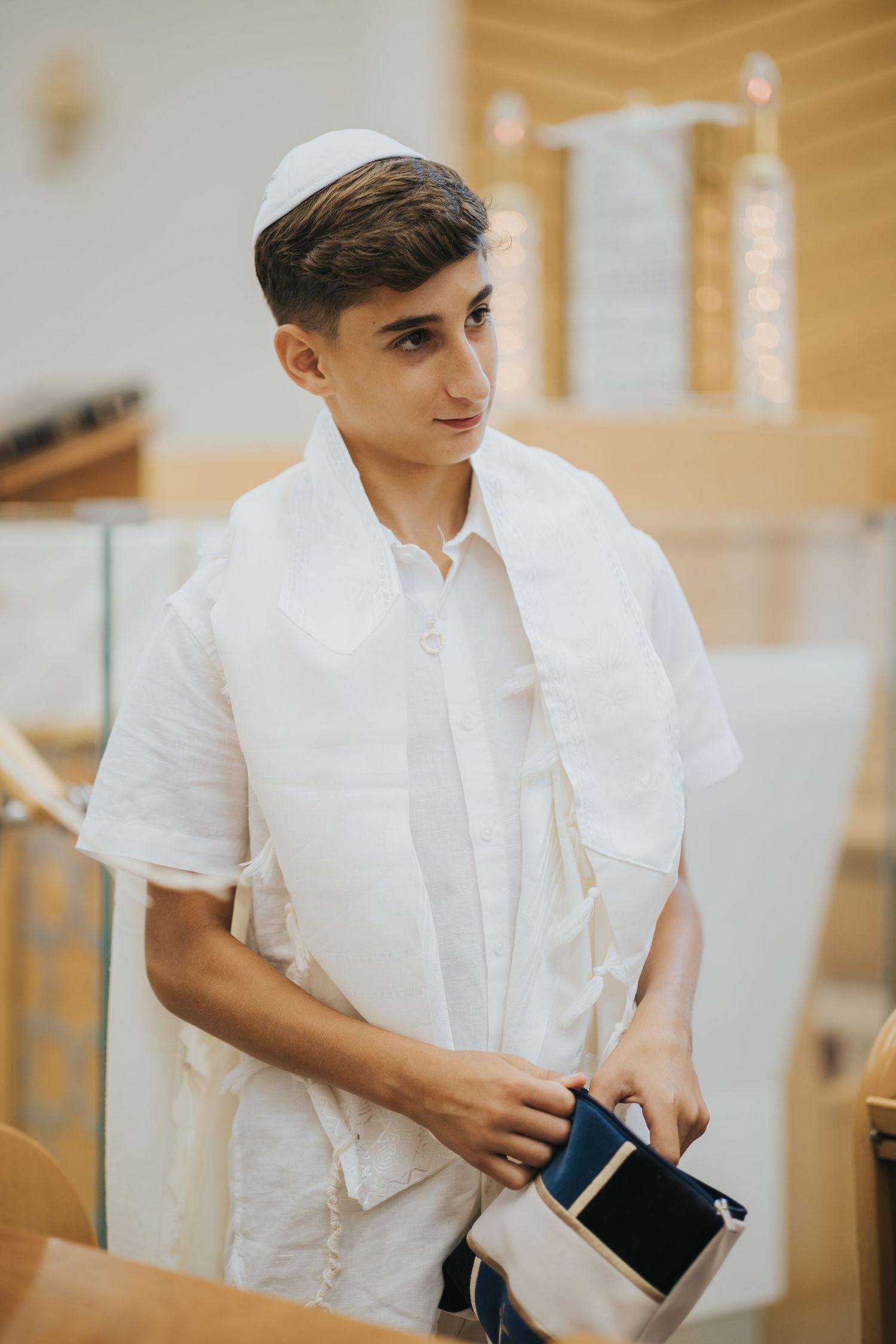 A person in white attire and kippah stands in a synagogue setting.