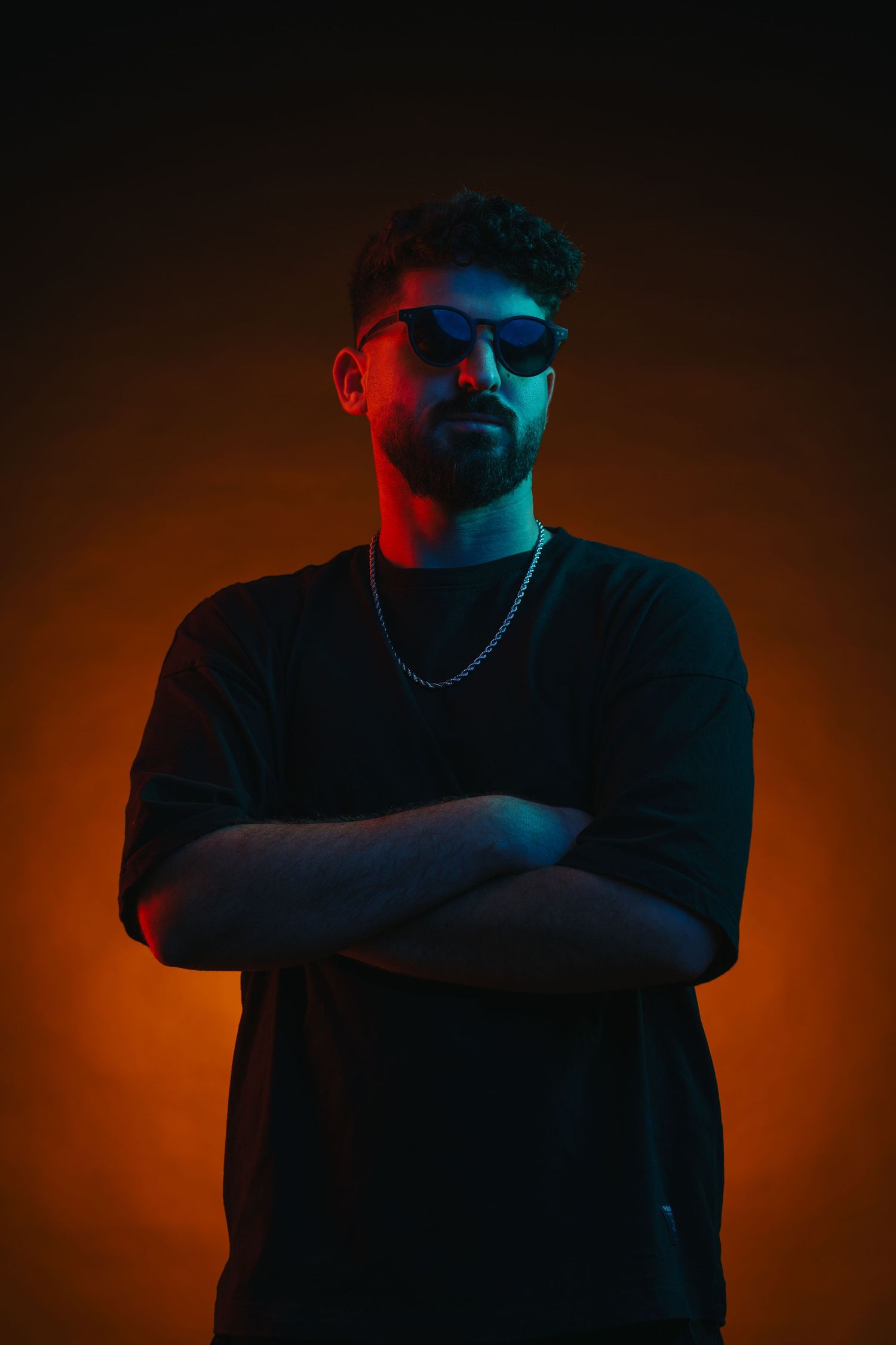 Dramatic portrait with orange and blue lighting featuring sunglasses and crossed arms pose.