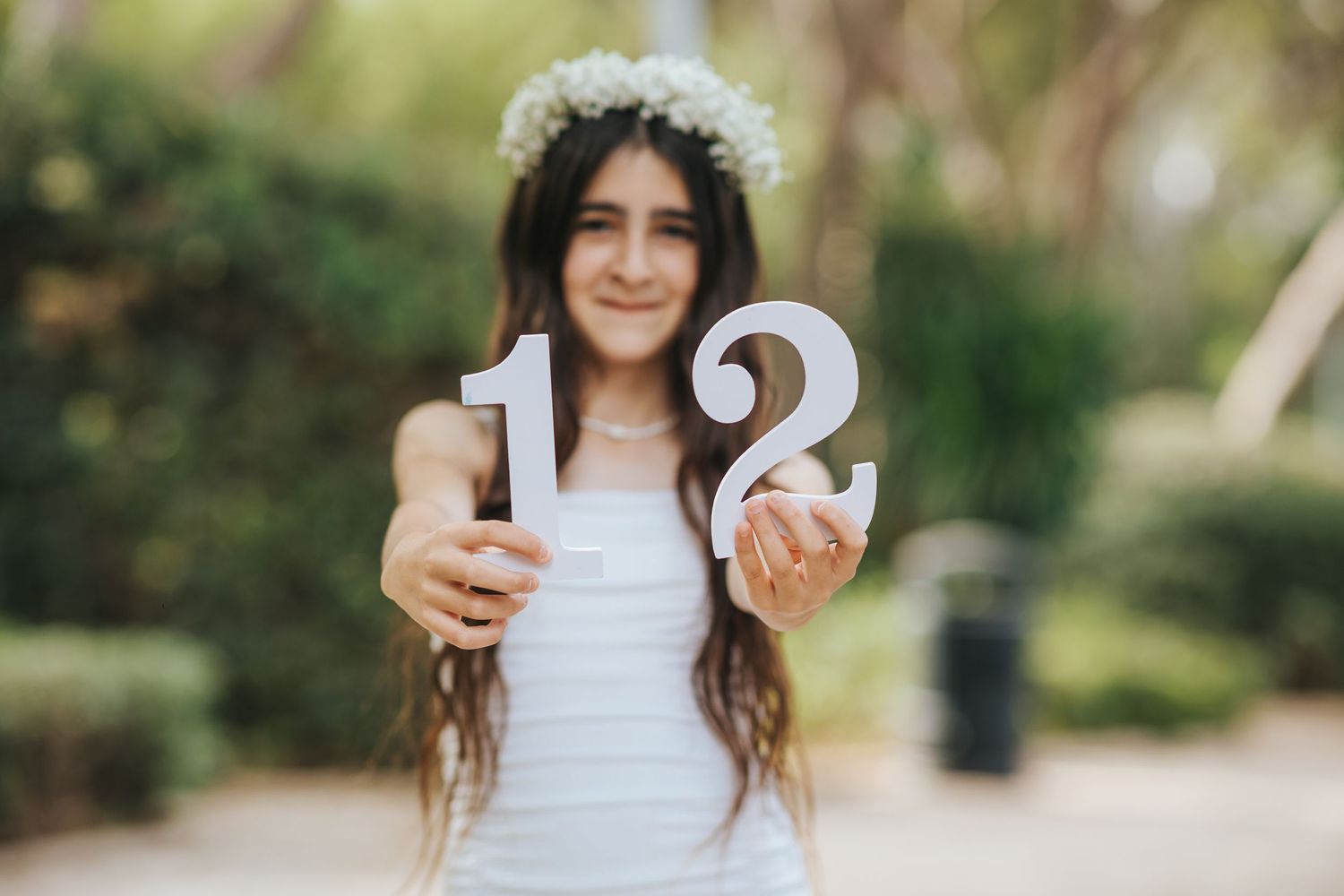 Person in white dress and floral crown holding number 12 numerals in outdoor garden setting.