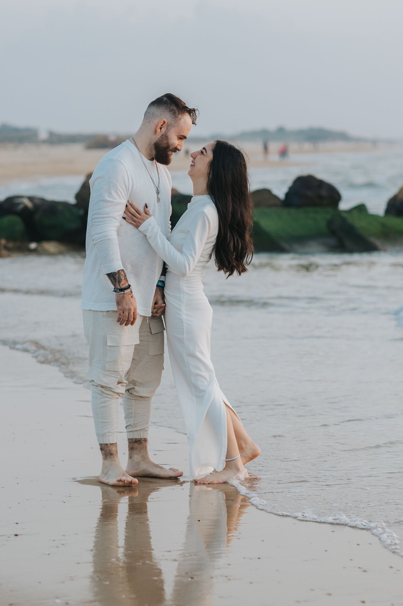 Romantic couple in white outfits sharing an intimate moment on a beach with crashing waves in the background.