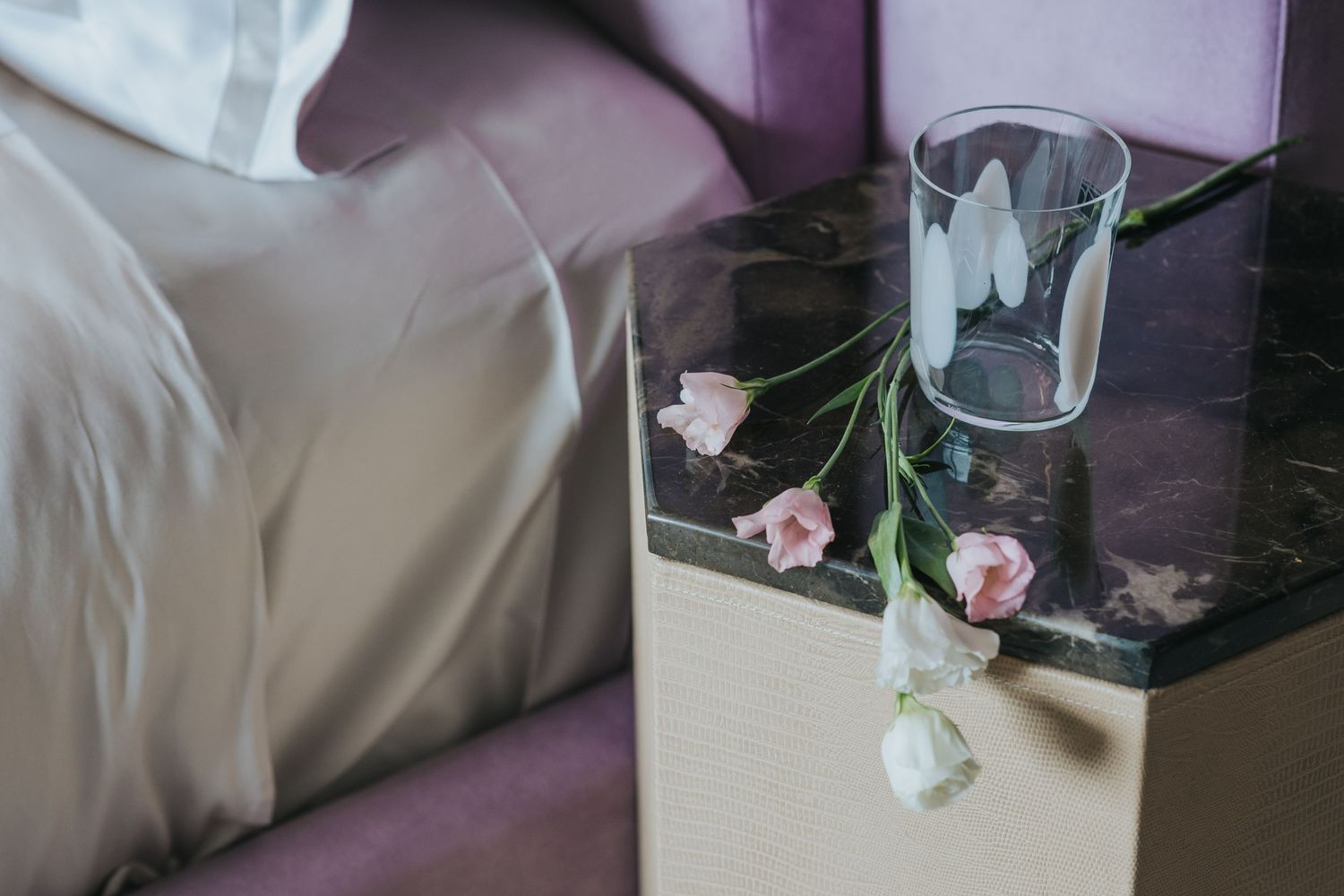 Delicate pink flowers in glass vase on bedside table with soft purple and white linens.