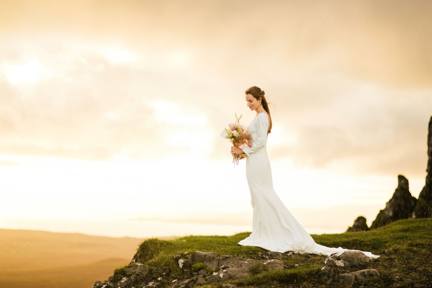 Bride holding bouquet on top of a cliff in Scottish Highlands' elopement at sunrise.