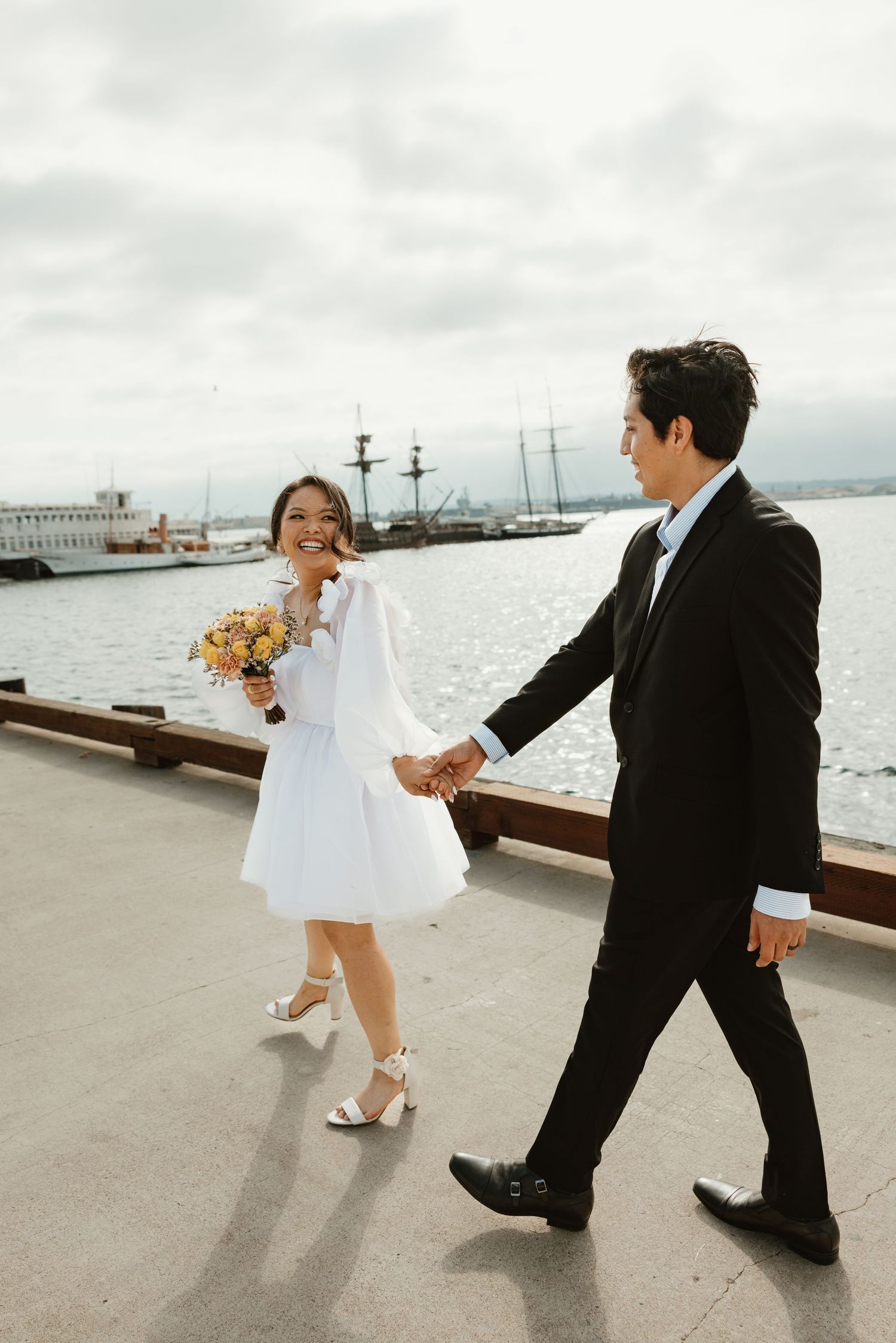 A couple in wedding attire walks playfully along a waterfront pier with city views in the background in Downtown San Diego.
