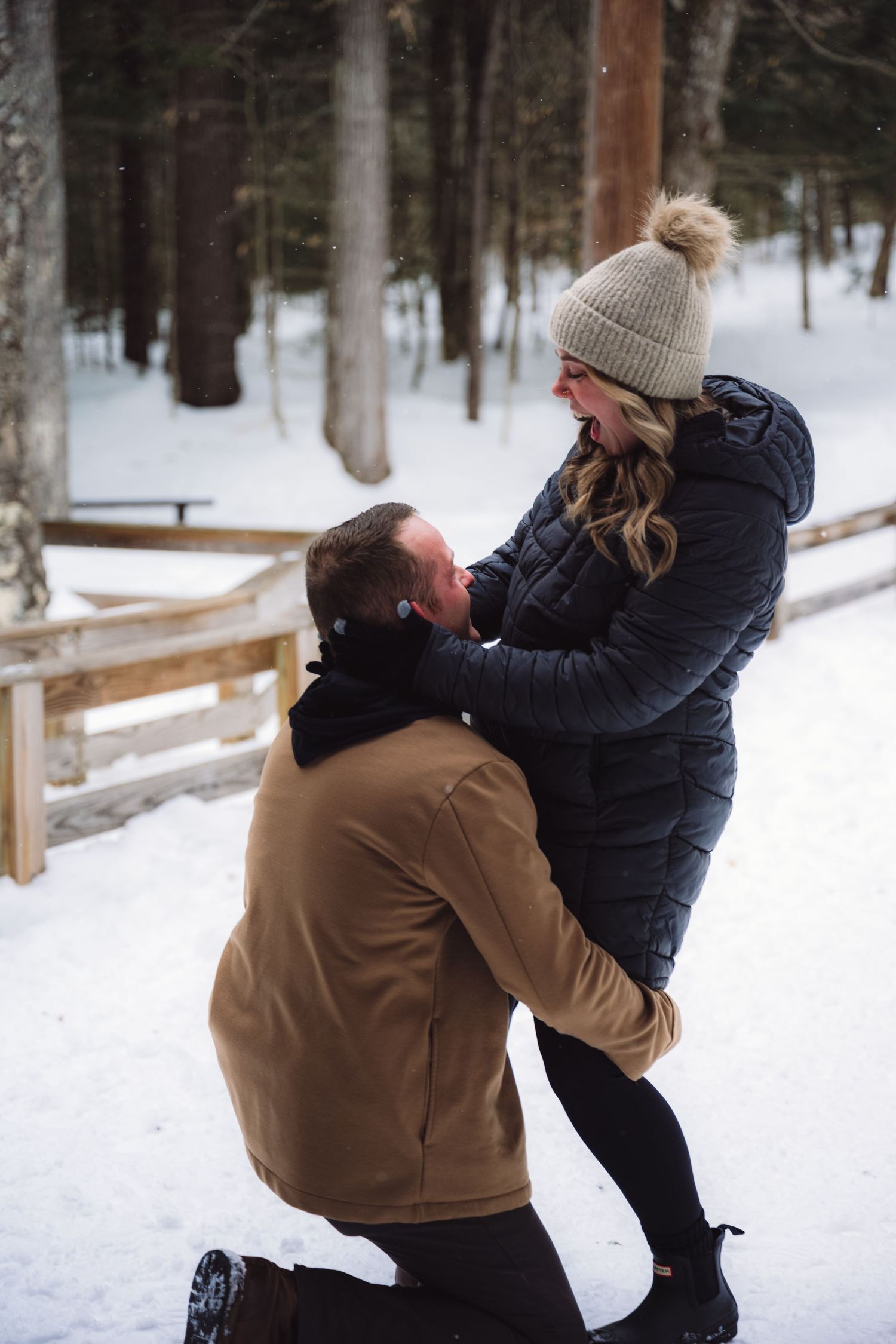 A couple embraces playfully in a snowy winter forest setting with evergreen trees in the background.