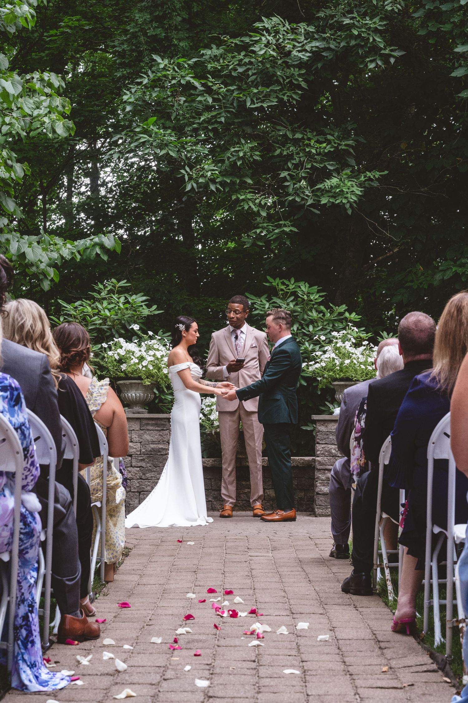 An outdoor garden wedding ceremony with rose petals scattered along a brick pathway.