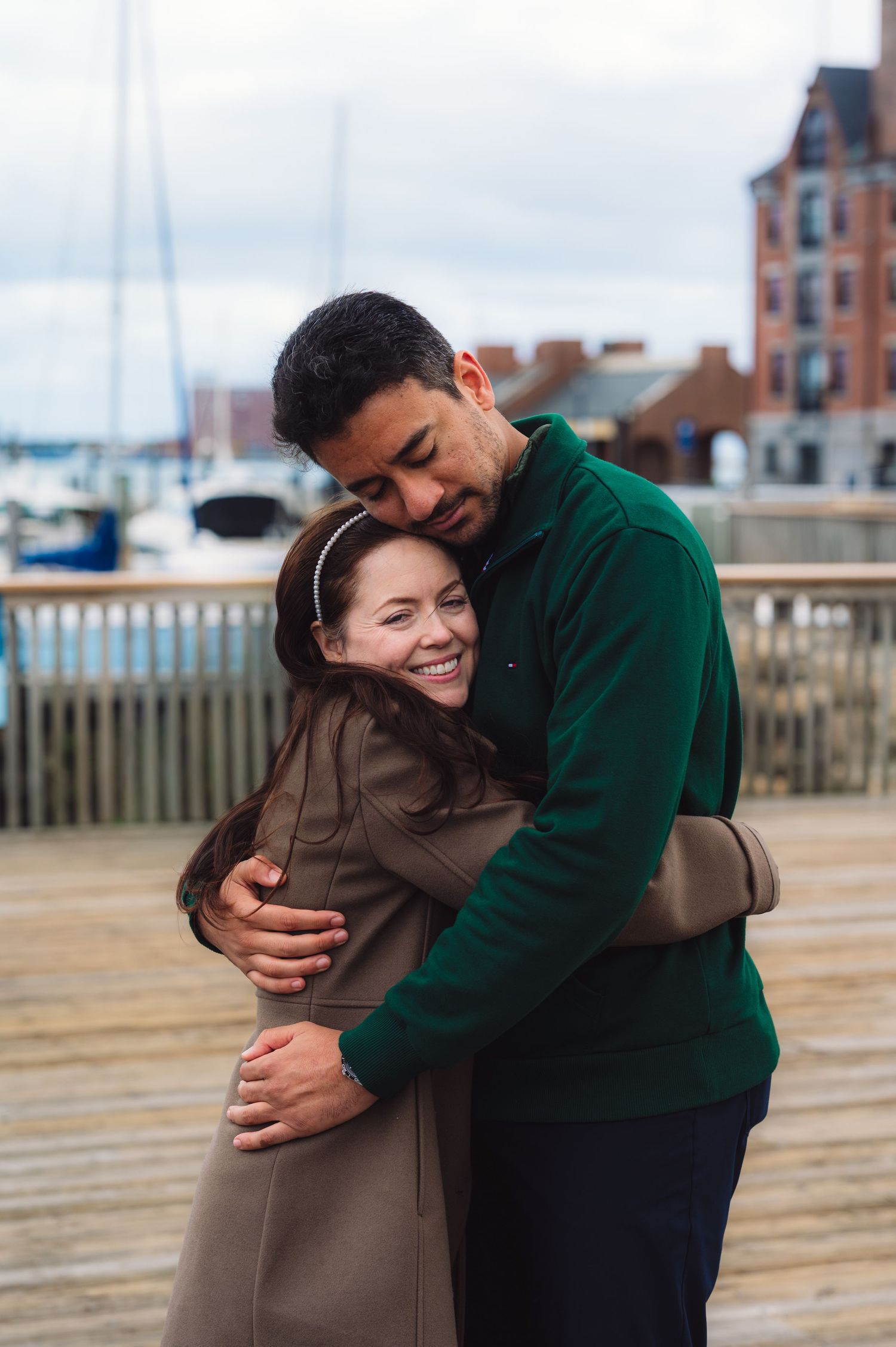 A couple embraces on a wooden boardwalk near a marina with boats and buildings in the background.