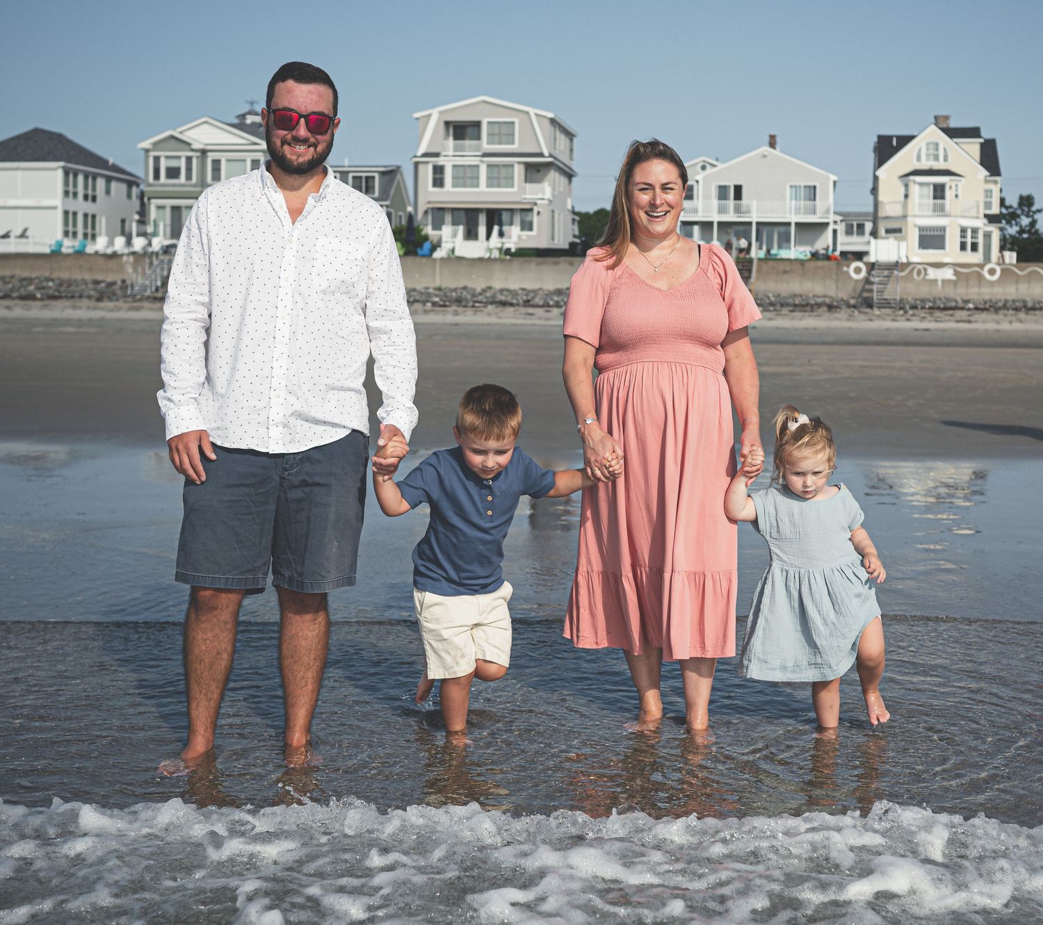 A family of four stands together at the edge of the ocean waves on a beach with coastal houses in the background.