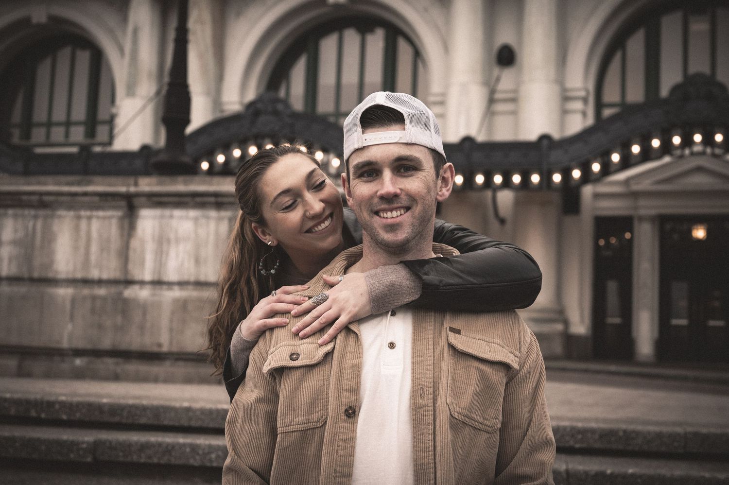 Couple poses for engagement photos in formal attire against stone architecture background.