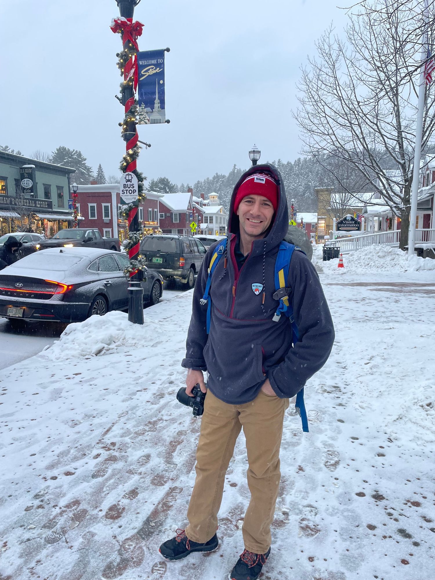 Person in winter clothing poses in snowy small town street with holiday decorations.