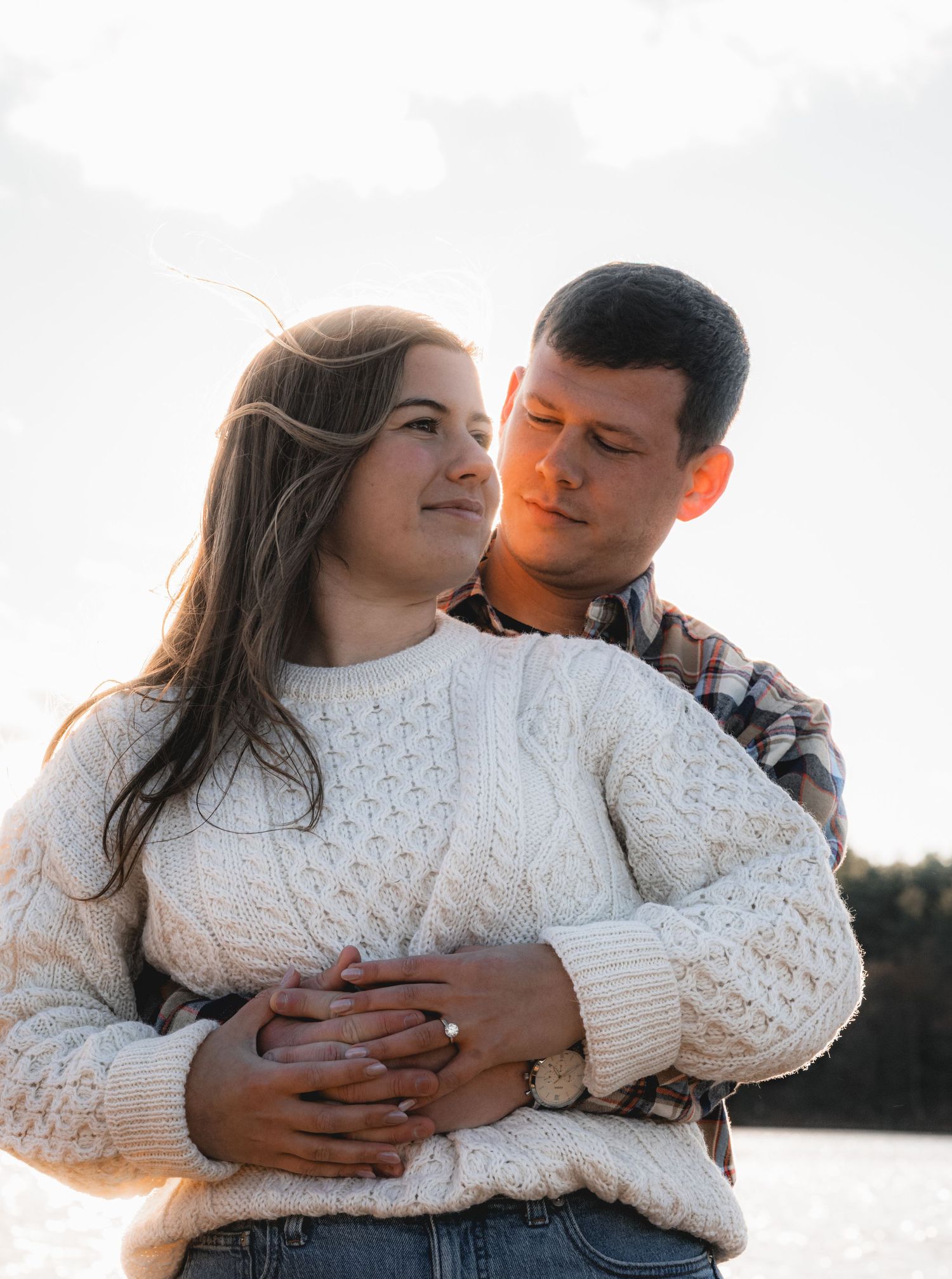 A couple in a white knit sweater and denim embrace outdoors during sunset by a lake.