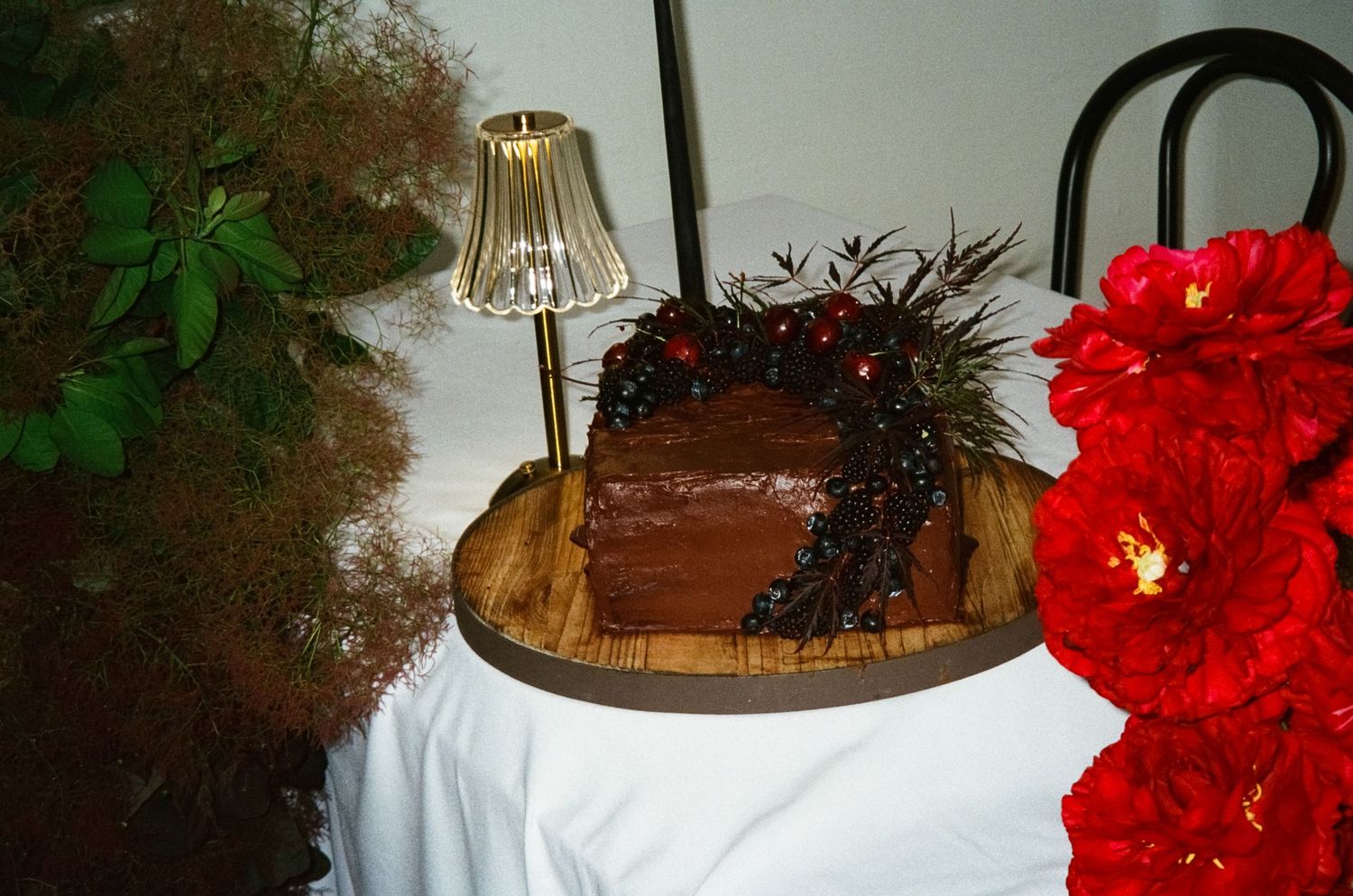 A round chocolate cake with dark chocolate drips on a wooden serving board next to a red floral decoration and lamp.