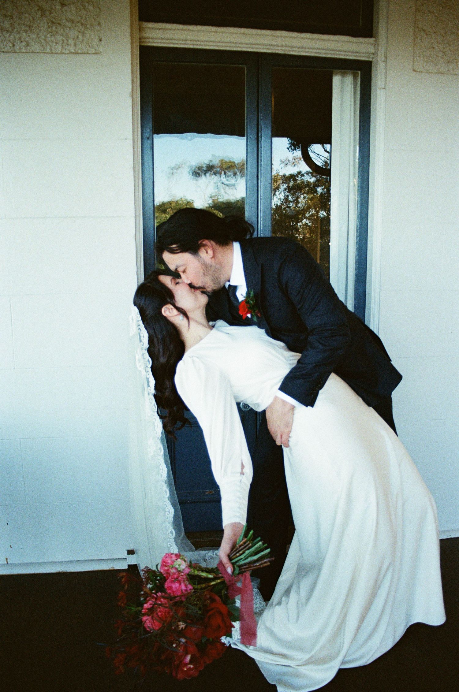 A romantic wedding photo showing a traditional dip kiss pose with red flowers and a white dress against a window backdrop.