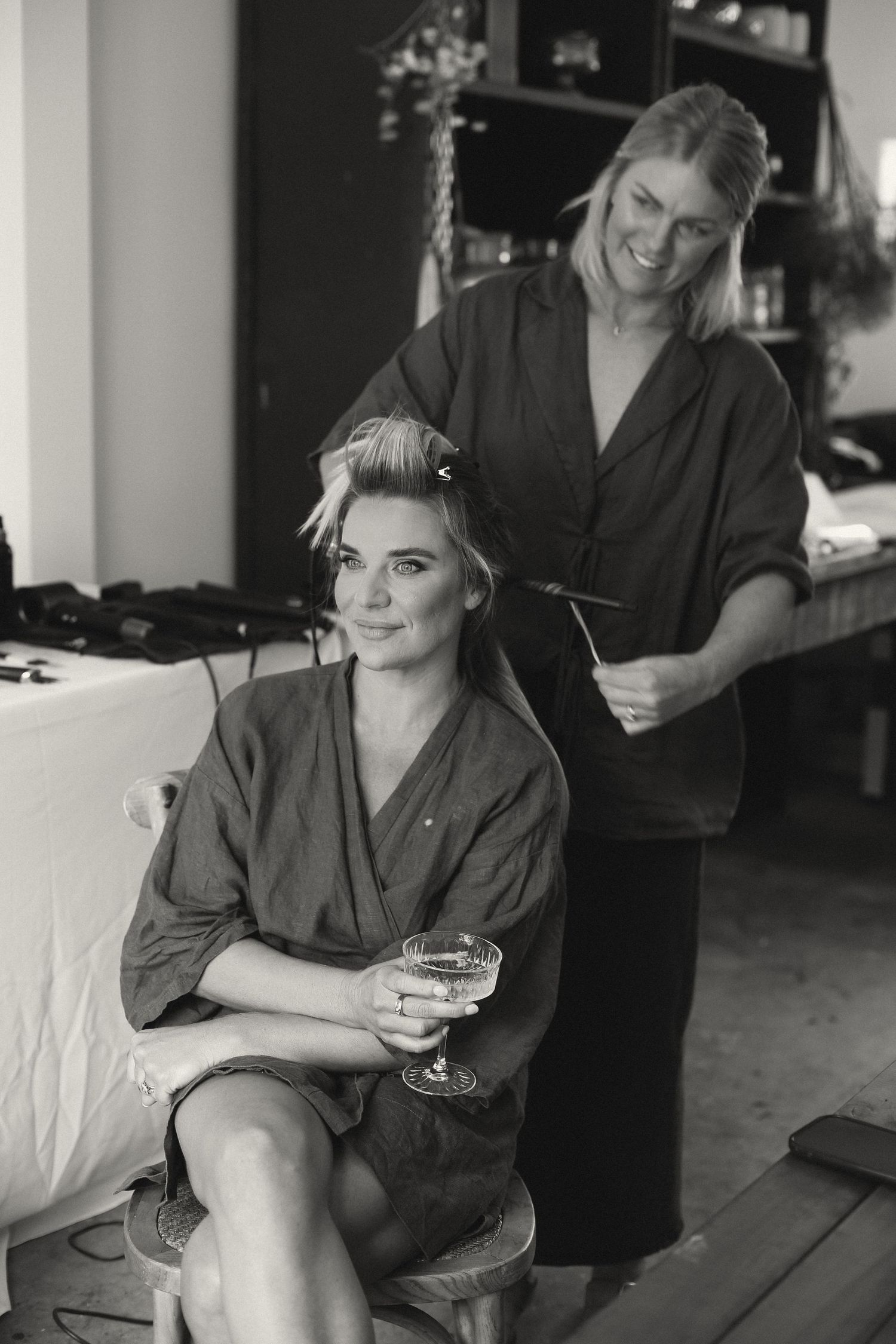 Black and white portrait of a person in a robe sitting on a couch holding a drink while having their hair styled.